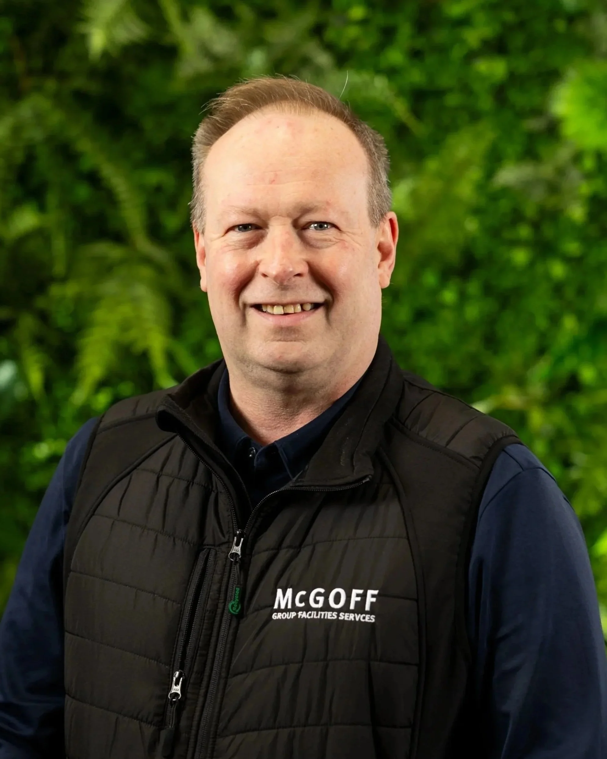 A smiling man with light skin, short light brown hair, and blue eyes, wearing a black quilted vest with the logo 'MCGOFF GROUP FACILITIES SERVICES' on it. He is outdoors with a lush green background of trees.