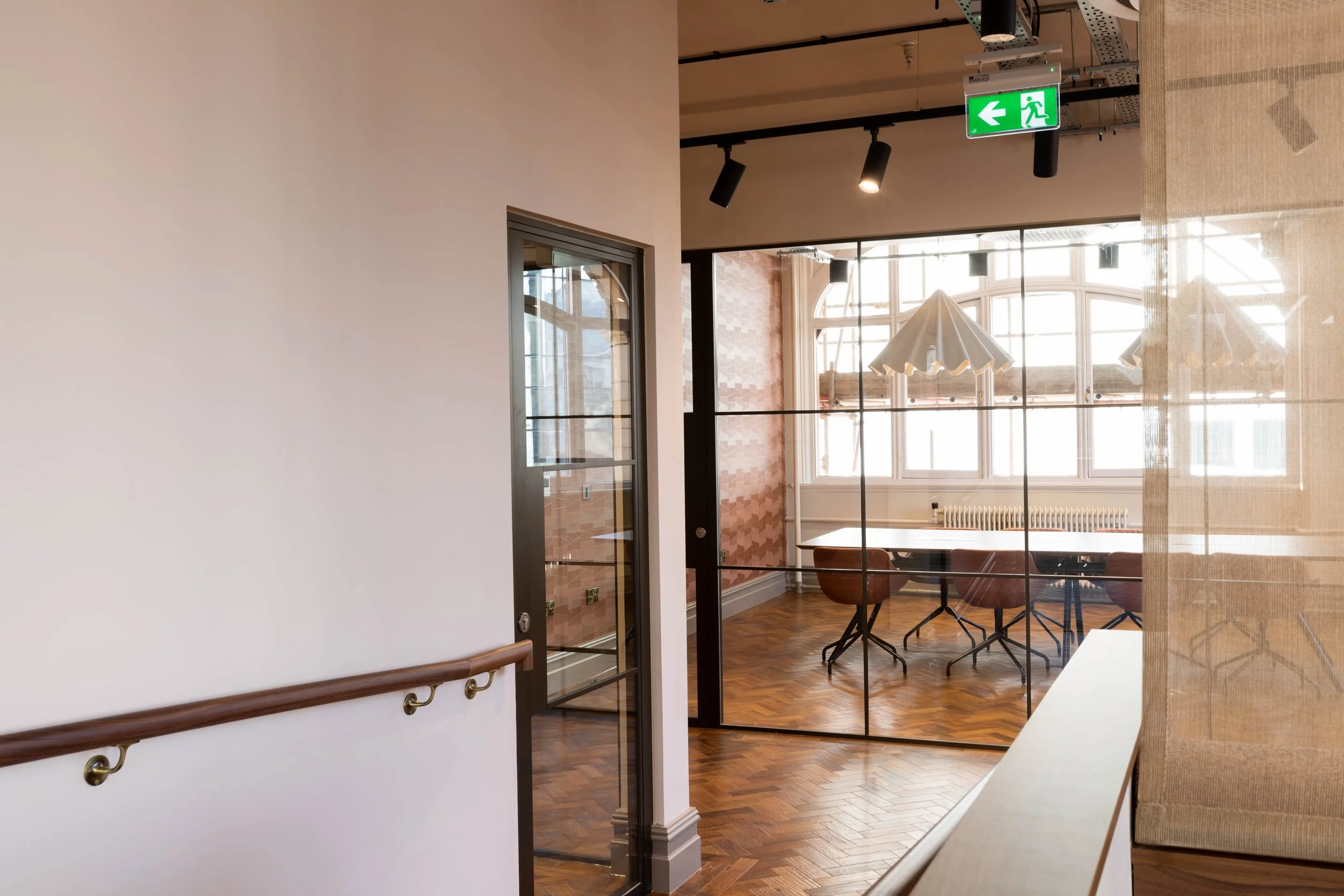 Interior of a modern building with large windows, a glass door, and a seating area with chairs and tables, illuminated by hanging lamps and natural light.