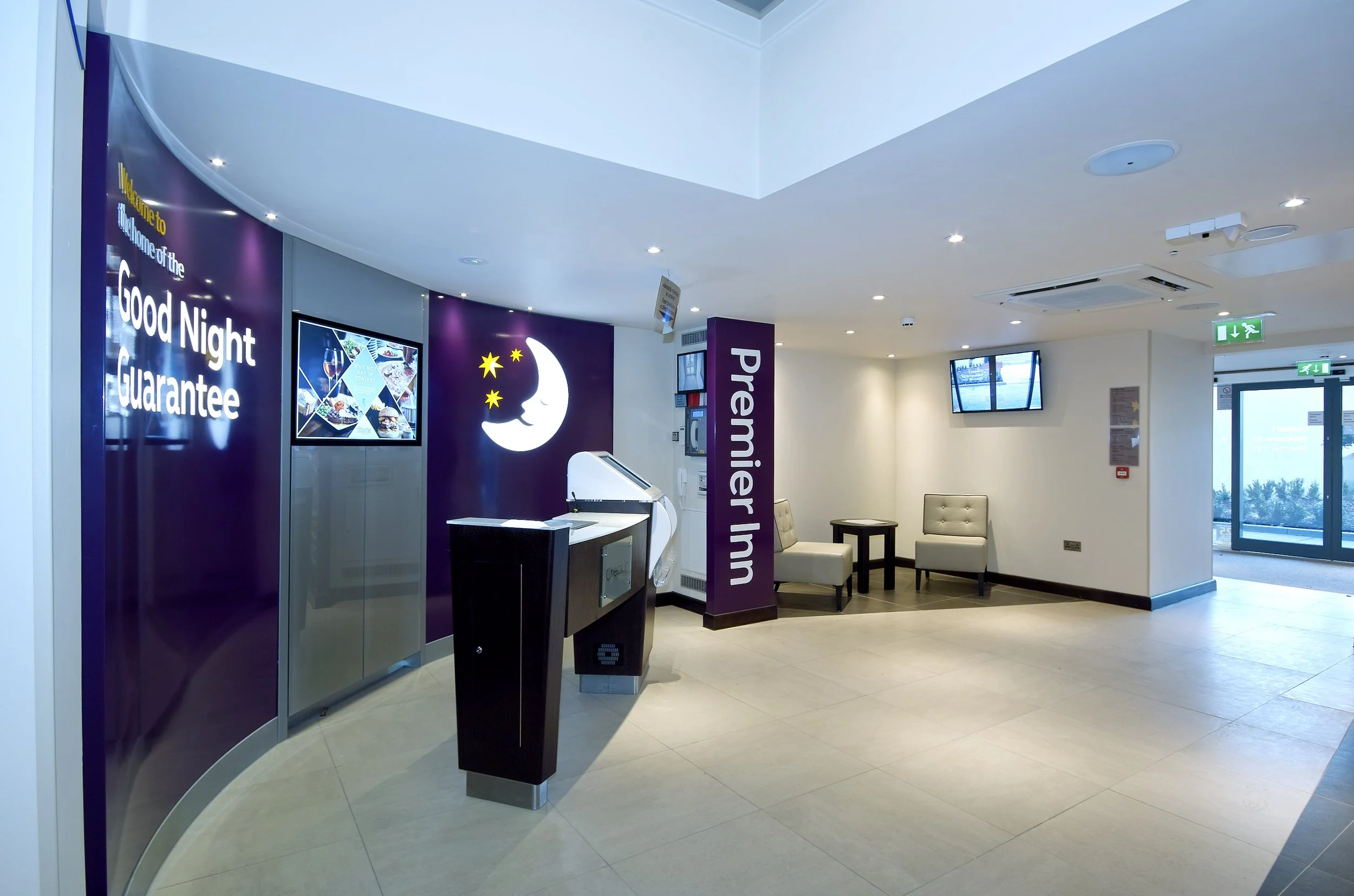 Interior of an airport or passenger terminal, showing seating area with chairs, digital screens, and signs indicating 'Premier In' and welcoming message with a moon and stars graphic.
