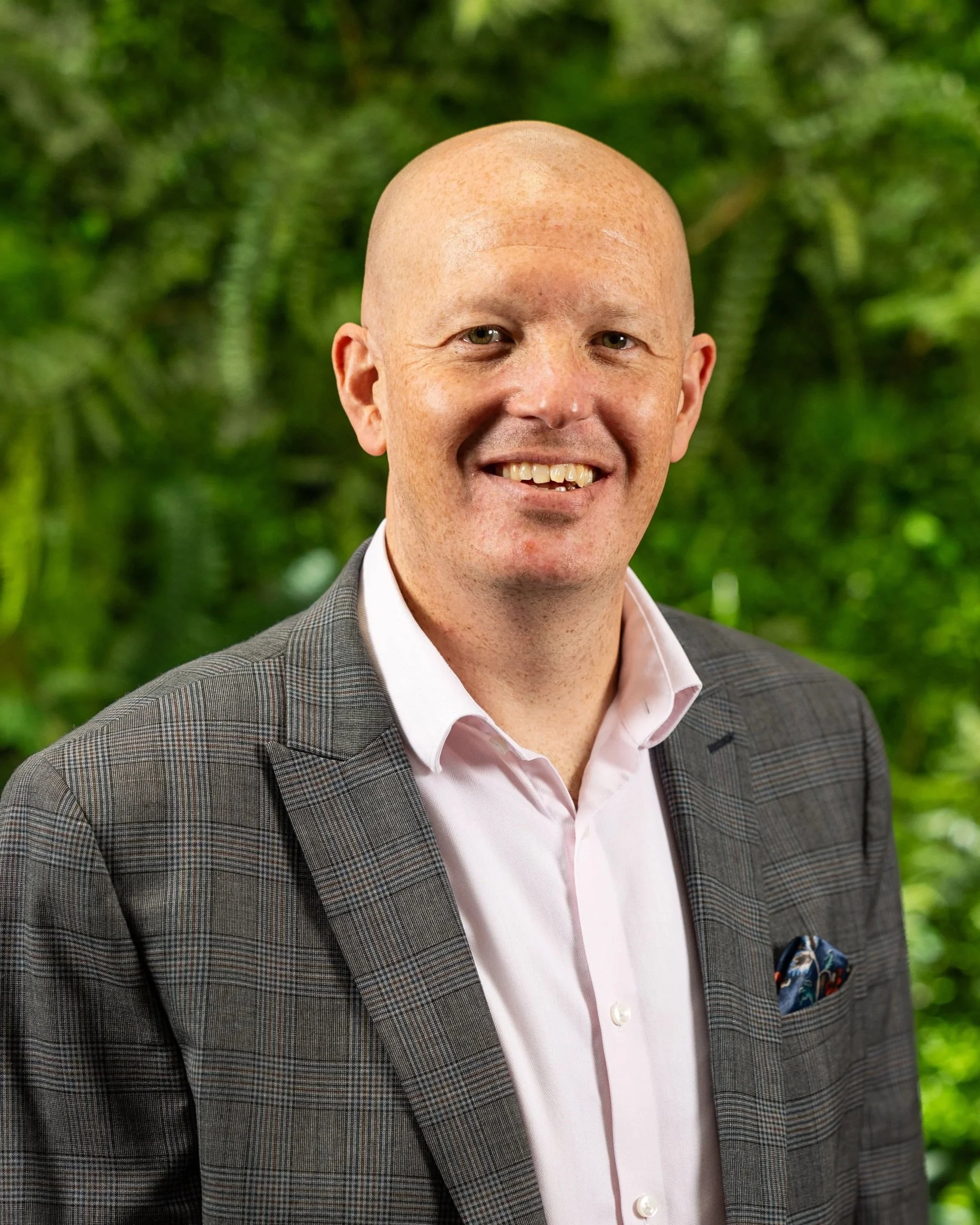 A smiling man with a bald head, dressed in a gray checkered suit, a pink shirt, and a colorful pocket square, standing outdoors with a lush green background.