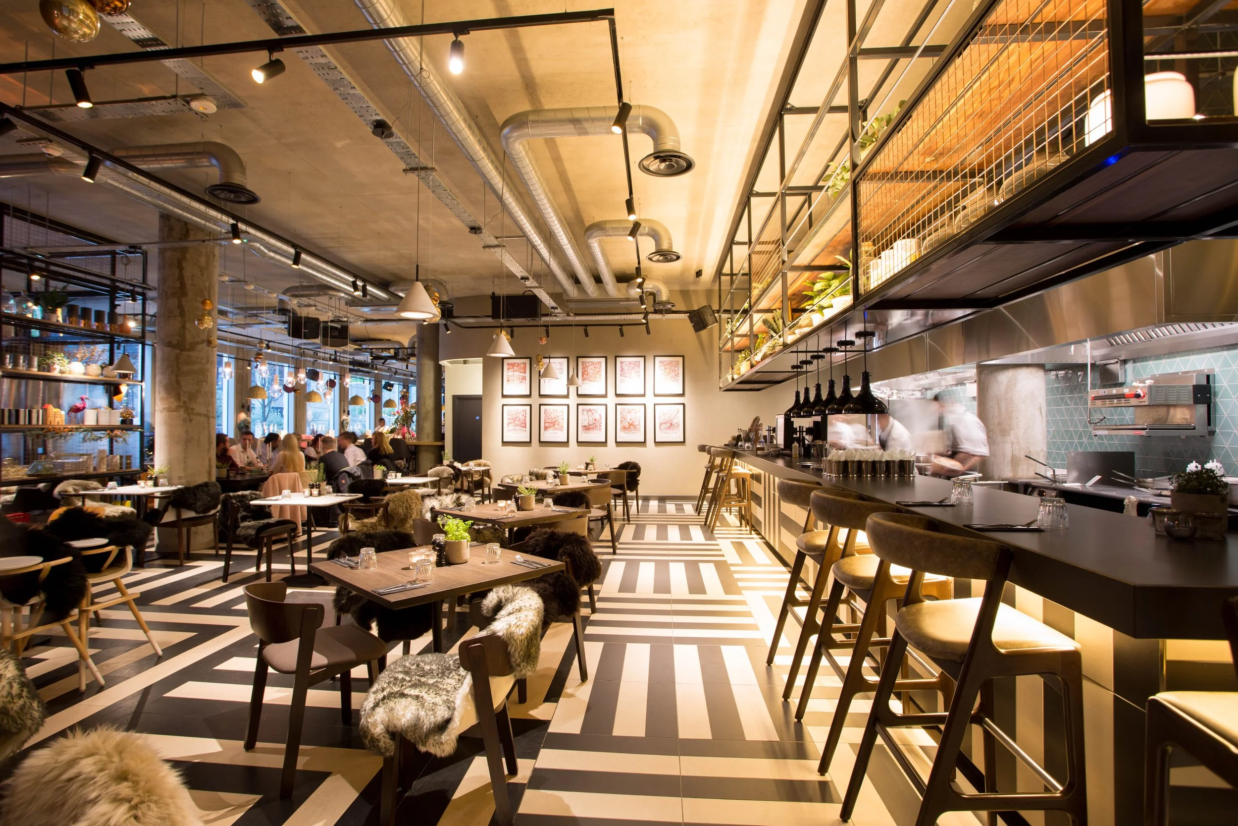 Interior of a modern restaurant with black and white striped flooring, wooden tables, and chairs with faux fur seat covers. The ceiling has exposed ductwork and track lighting. There are hanging pendant lights over the bar area on the right, where ch