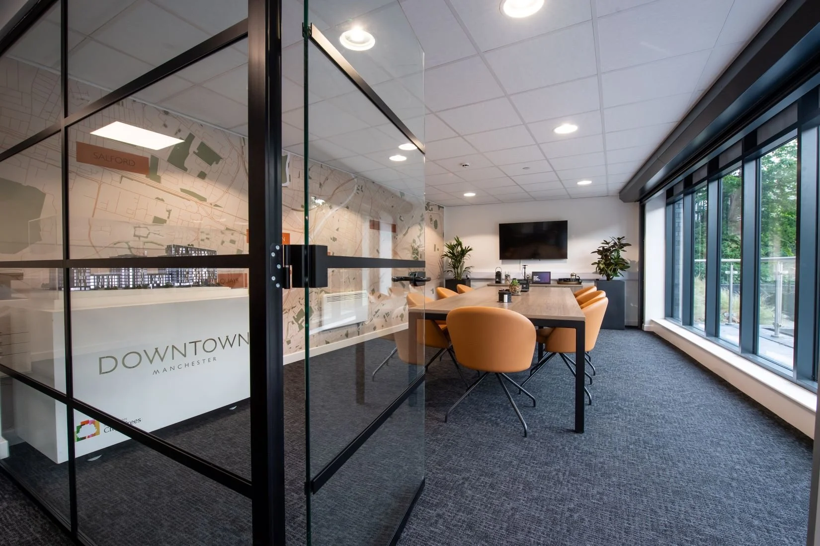 Modern conference room with a large wooden table, tan chairs, a glass wall with a city map graphic, a large window with greenery outside, and a TV on the wall.