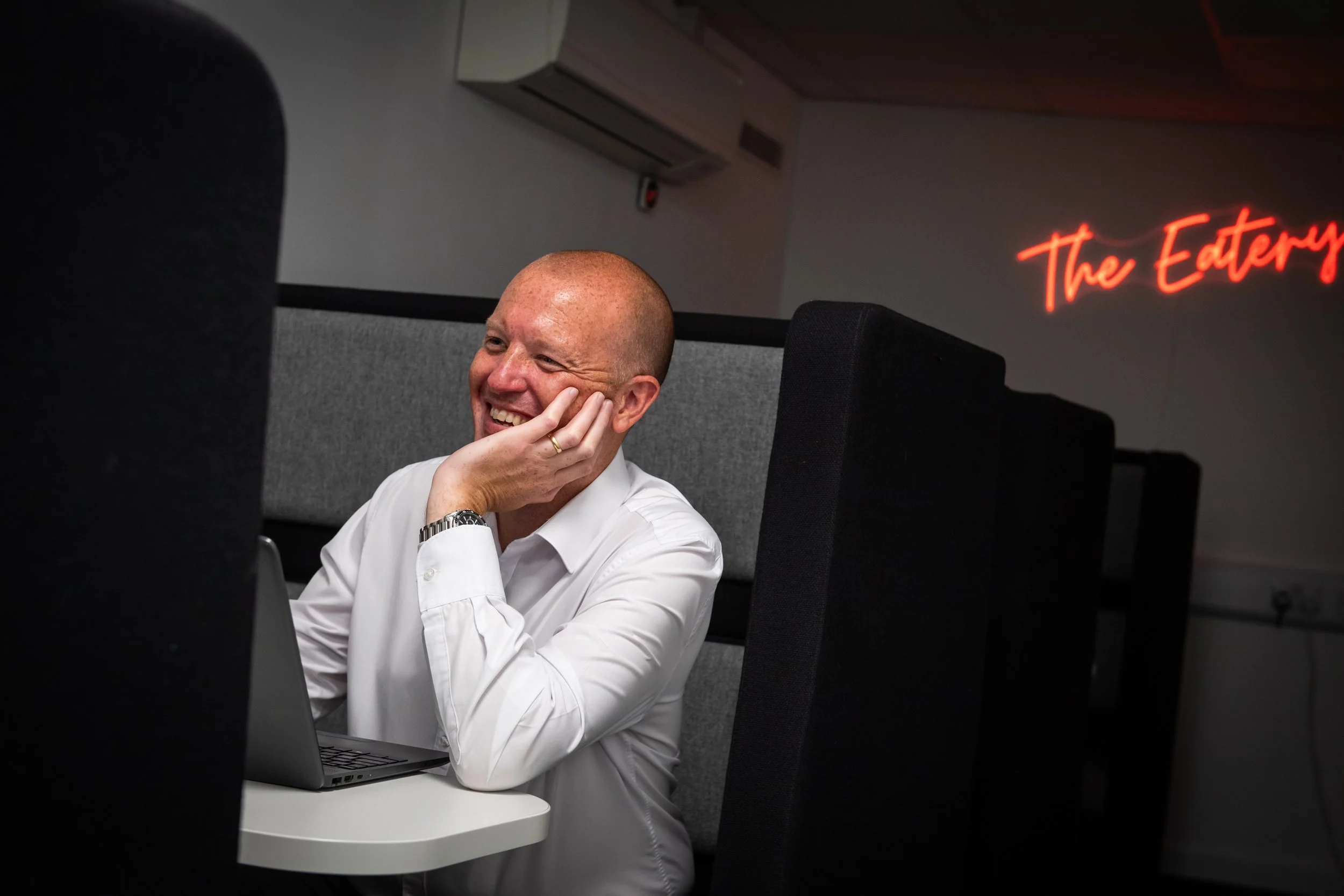 A man with a shaved head wearing a white shirt and a watch, smiling while sitting at a desk with a laptop. Behind him, there is a gray partition, a wall with a neon sign reading 'The Edge', and an air conditioning unit.