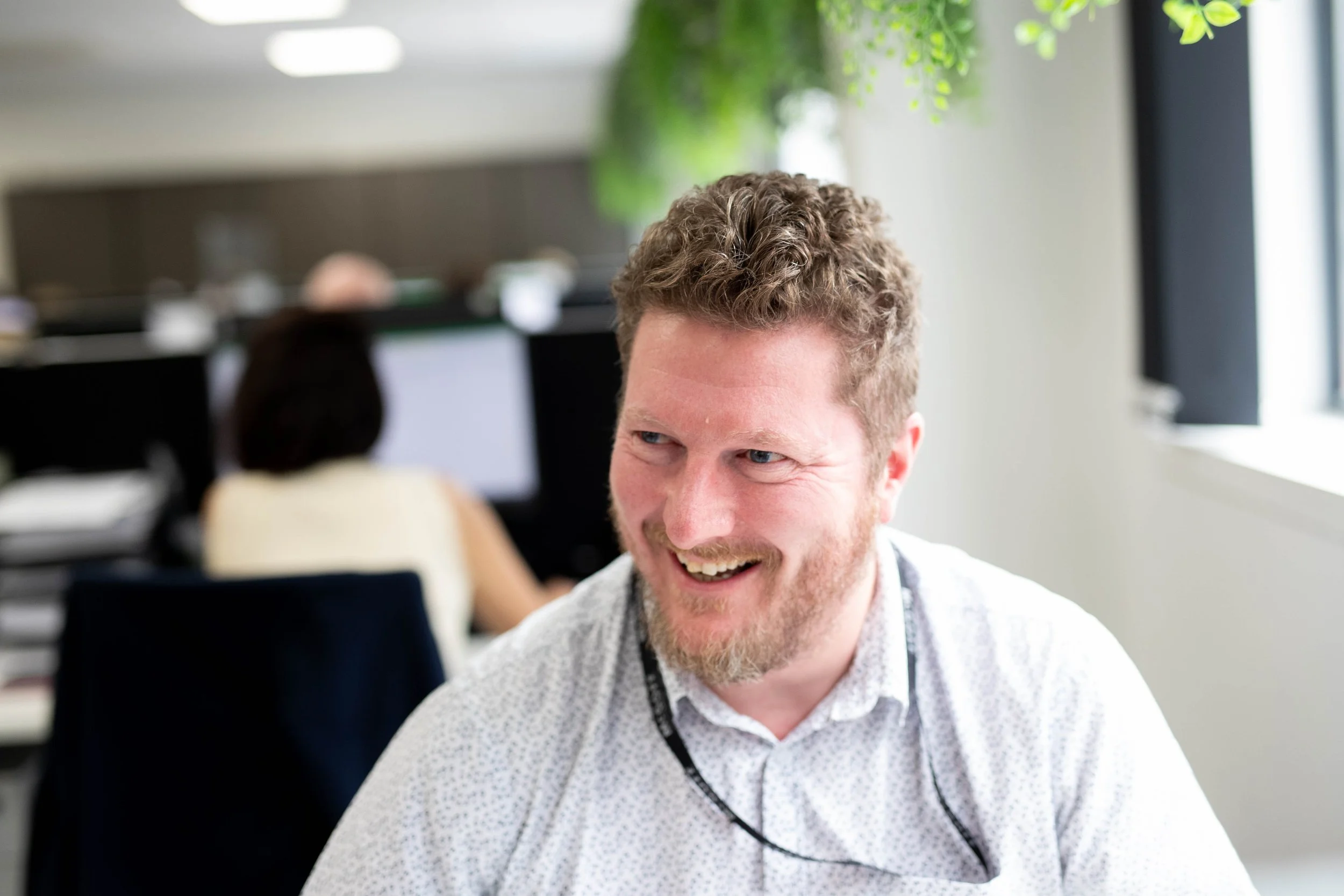 A smiling man with curly hair and a beard, wearing a white patterned shirt, in an office environment with blurred computer monitors and green plants in the background.