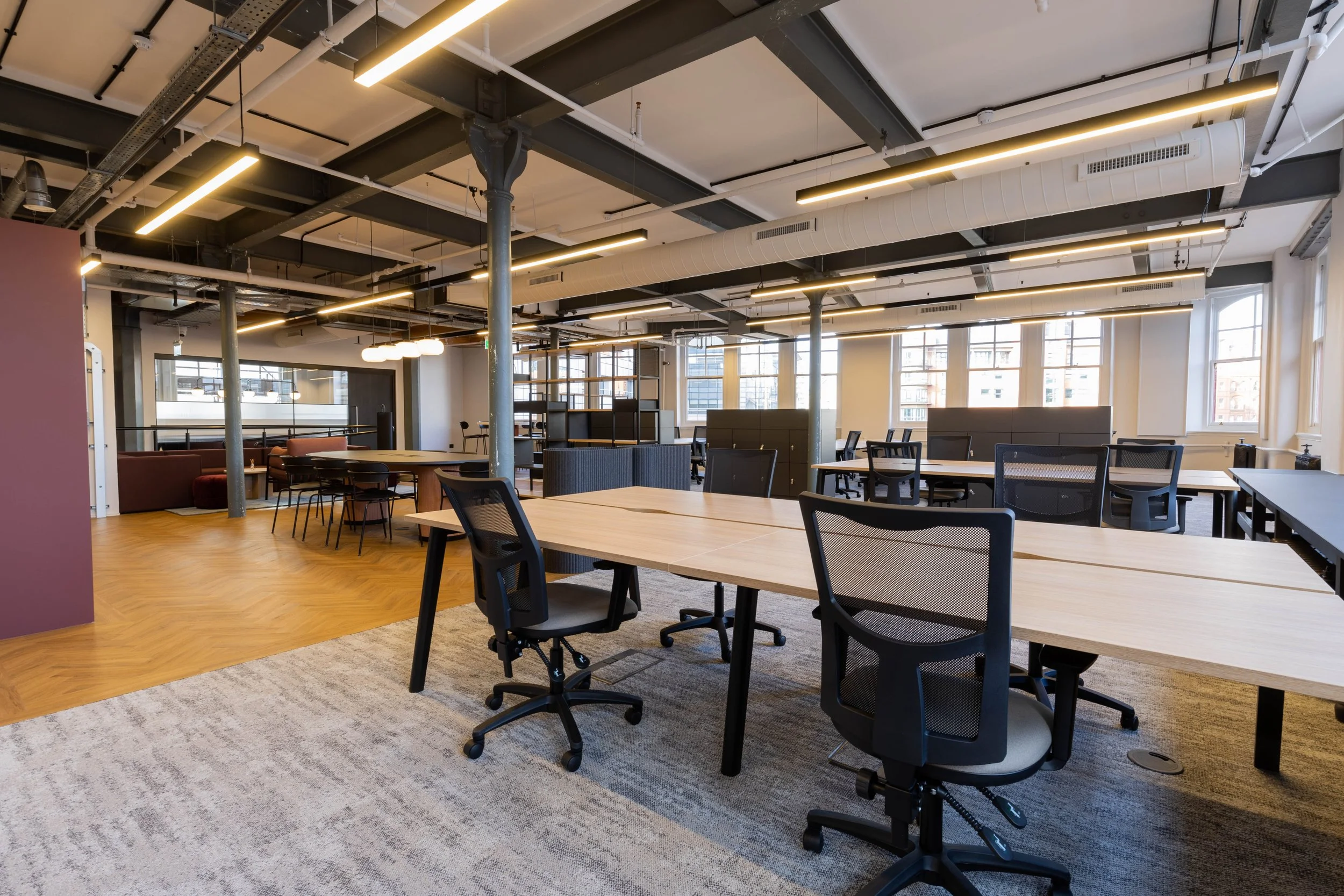 An open office space with high ceilings, exposed pipes, and large windows. There are multiple wooden desks with black office chairs, some of which are on wheels. The floor has a mix of hardwood and carpeted areas, and there are pendant lights and lon