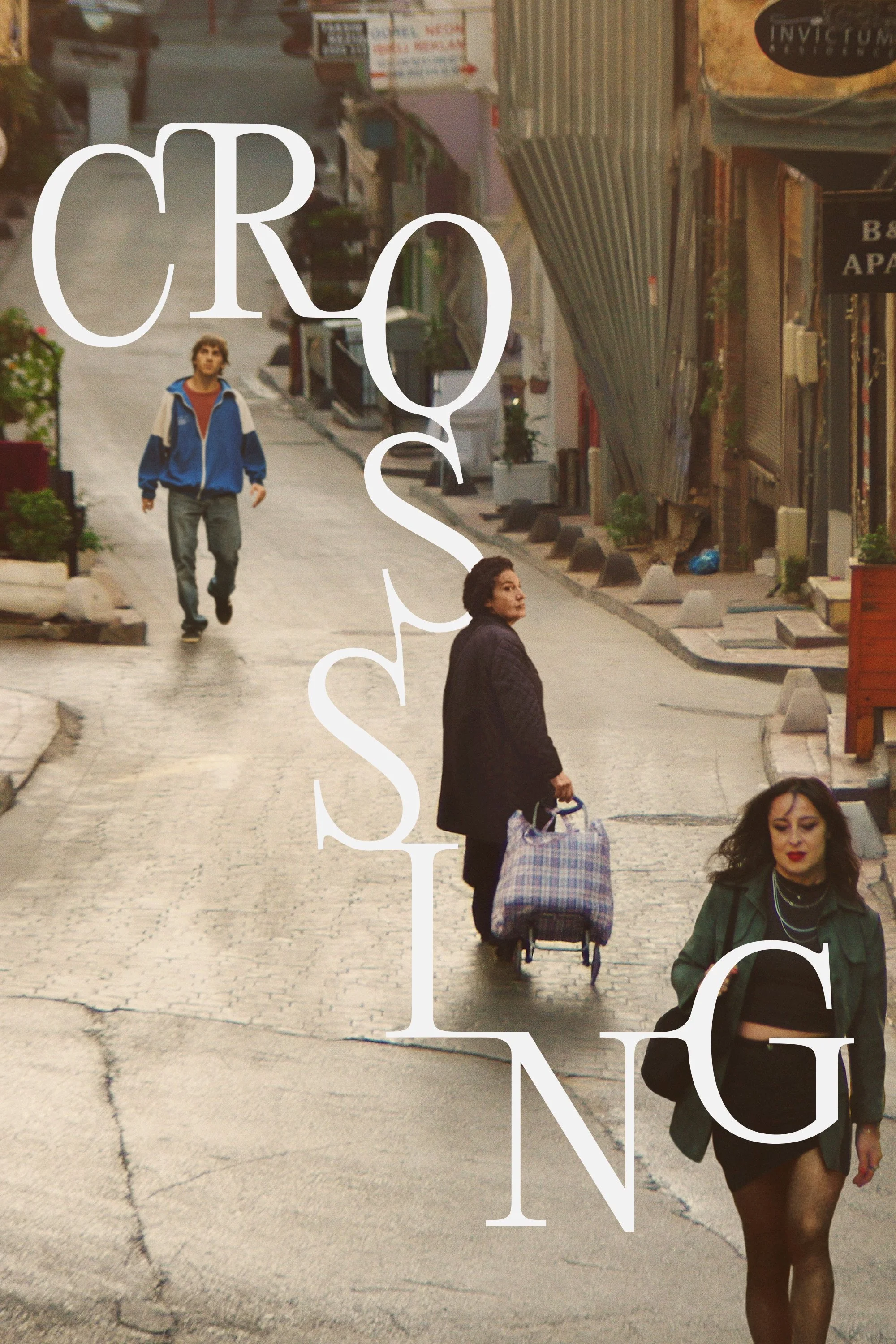 A street scene with several people walking. There is a young man in a blue jacket walking toward the camera, a woman with a shopping cart, and a woman in black clothing. The street is cobblestone, lined with plants and shop signs.