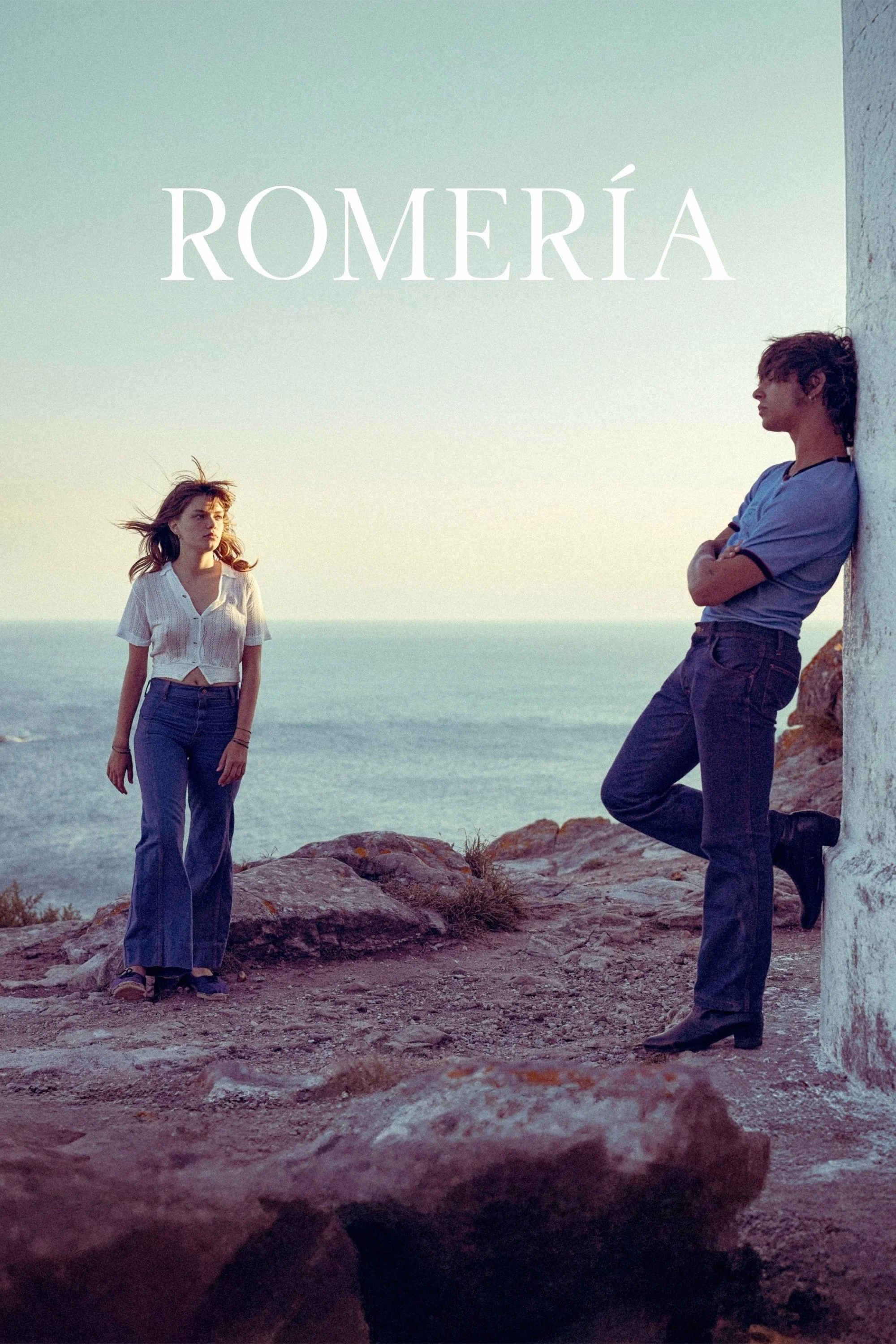 A young woman and man by the ocean at sunset, with the woman standing and the man leaning against a white wall with arms crossed.