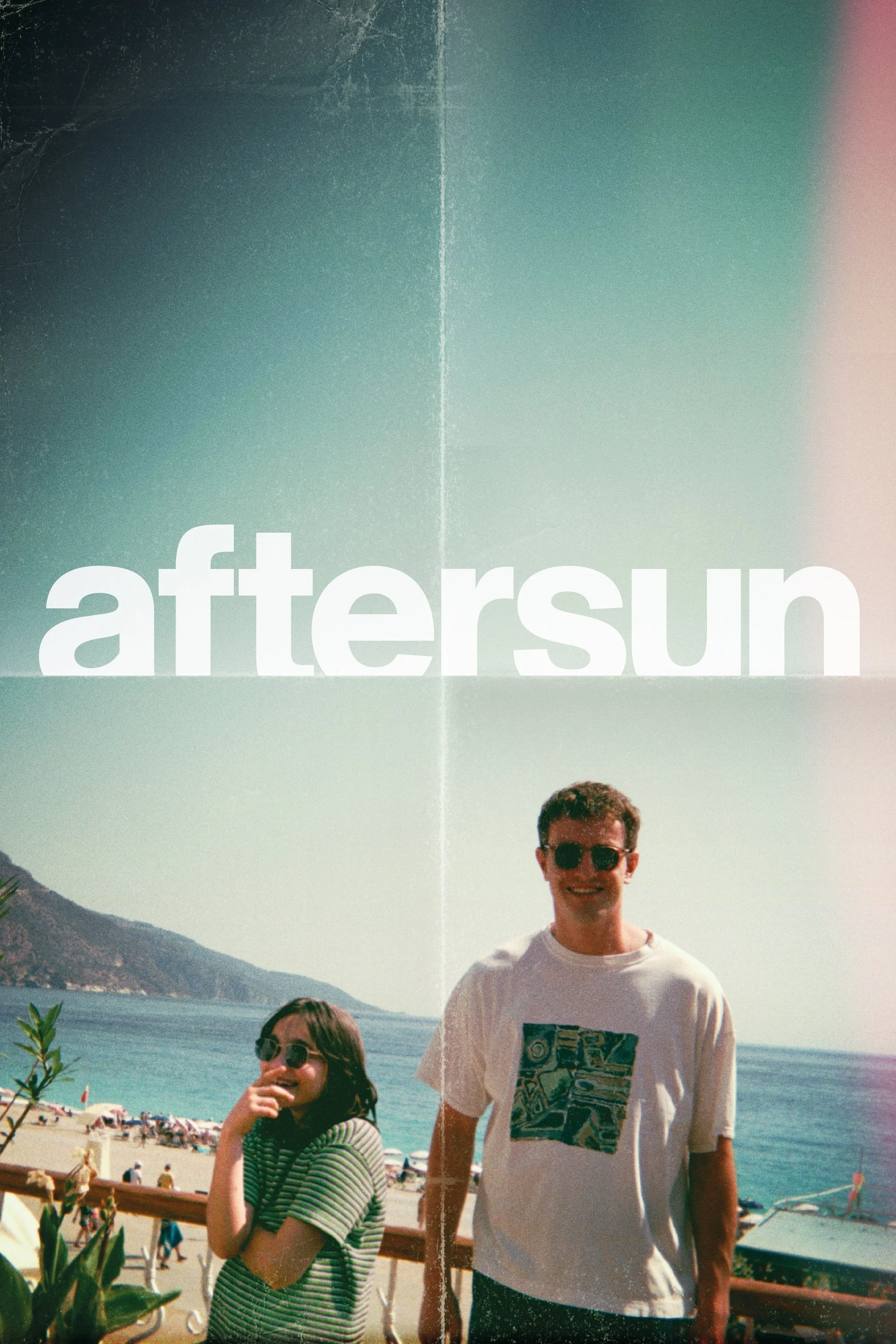 Two smiling people, a woman and a man, standing outside near a beach. The woman has long dark hair, glasses, and is wearing a striped shirt. The man has short dark hair, sunglasses, and is wearing a graphic t-shirt. They are in front of a scenic backdrop with water, mountains, and a clear sky, with the word 'aftersun' overlayed.