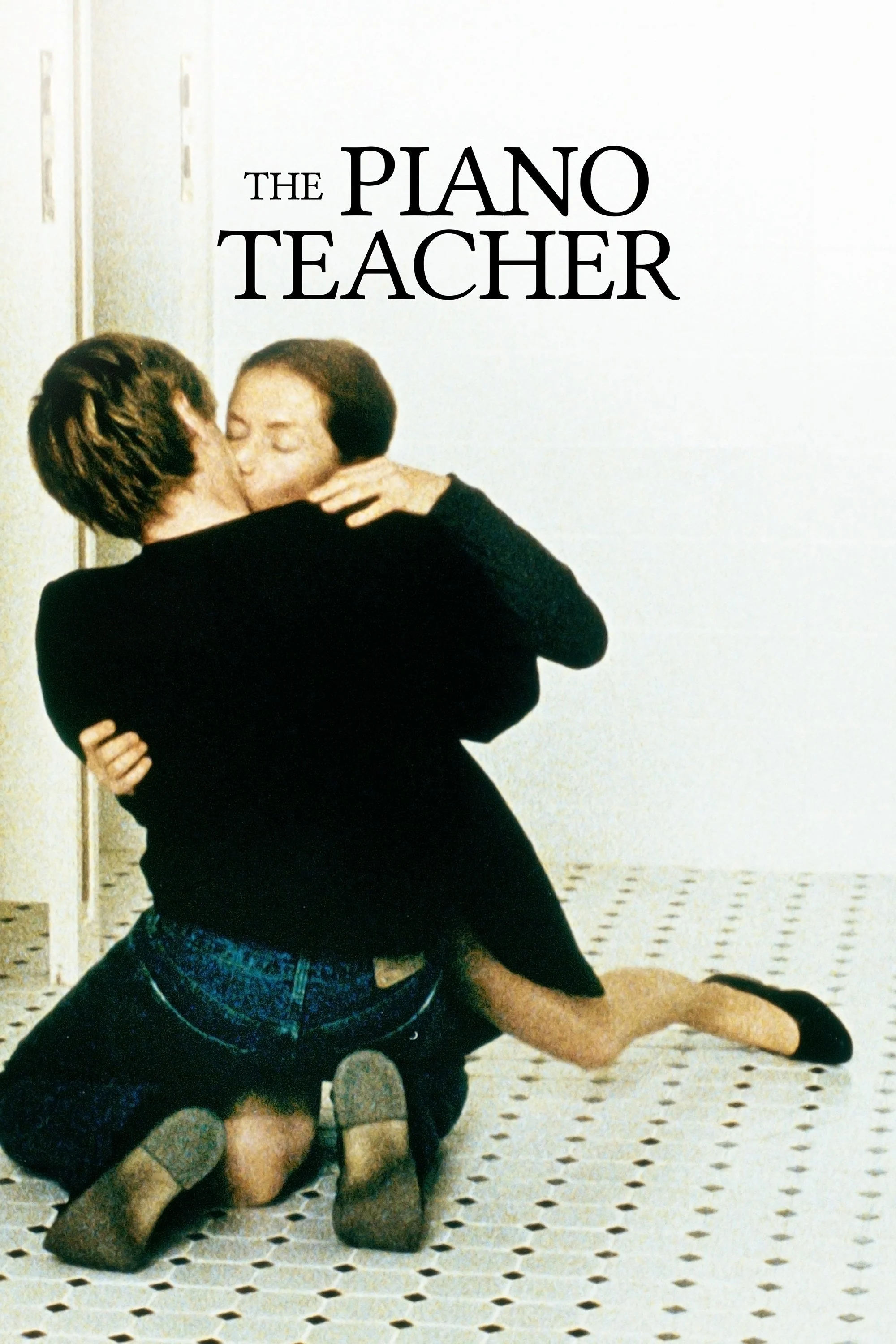 Two women are kissing while one is sitting on the floor and the other is kneeling. The woman on the floor is wearing a black top and denim shorts, while the woman kneeling is dressed in a black dress and black shoes. They are in a room with white walls and a patterned floor.