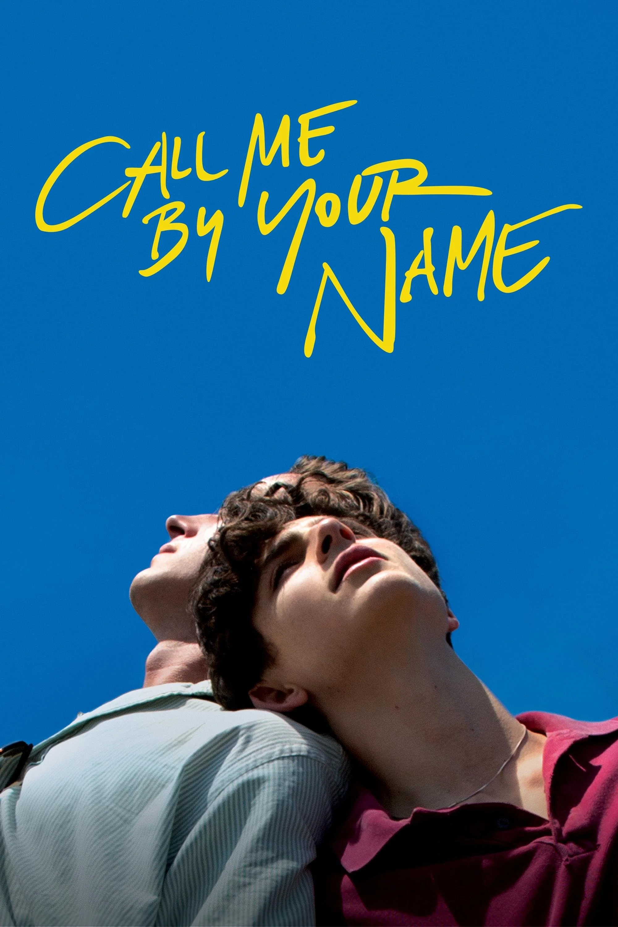 Two young men with dark hair and light skin, one in a white striped shirt and the other in a maroon shirt, standing close together outdoors under a bright blue sky. Text in yellow handwritten font above them reads "Call Me By Your Name."
