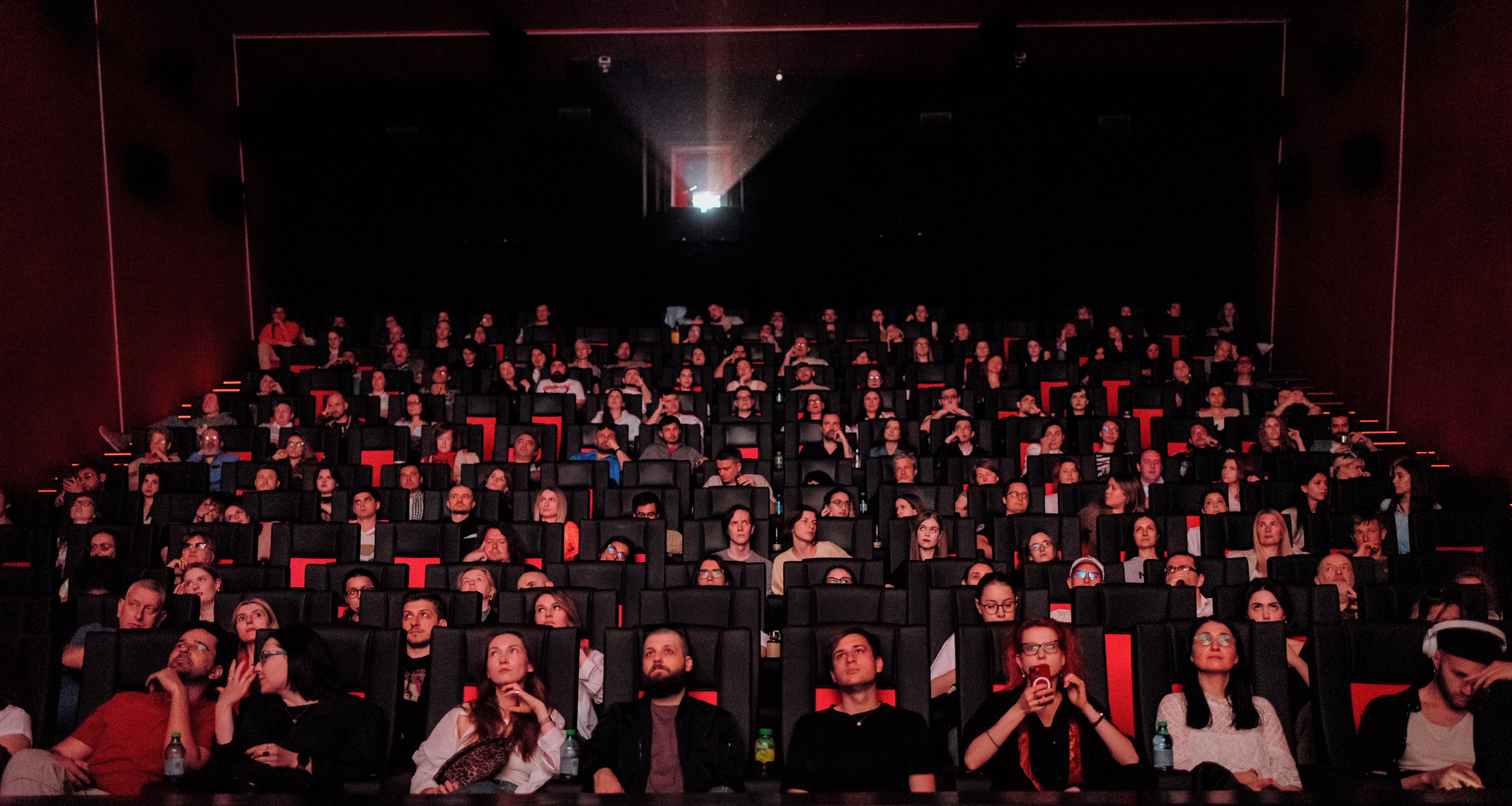 People sitting in a darkened auditorium watching a presentation or film, with some looking at their phones and others attentively awaiting the event.