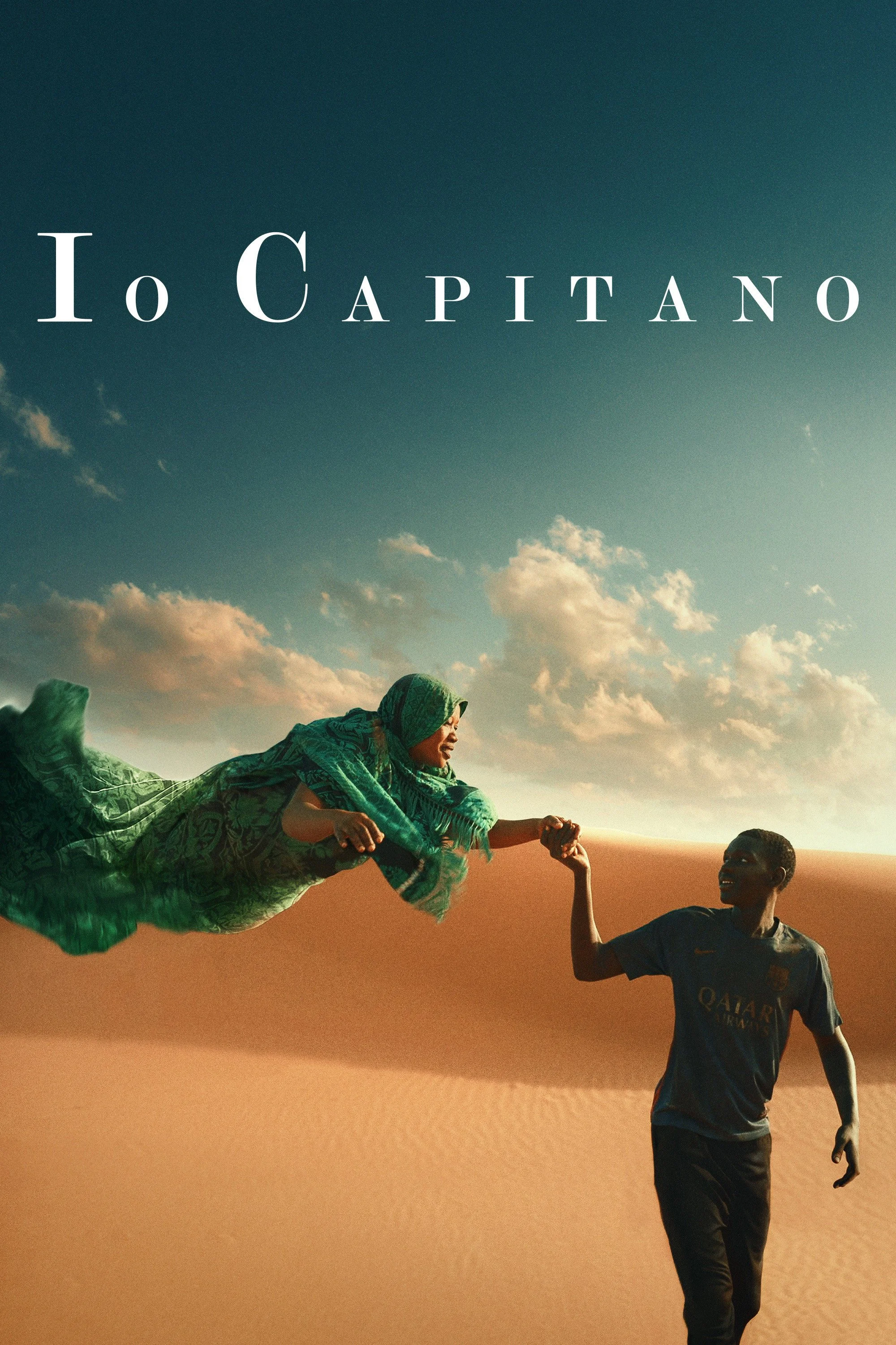 Two people in a desert, one floating horizontally wearing a green patterned outfit and scarf, the other standing and holding the floating person's hand, under a partly cloudy sky.