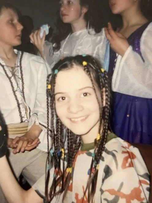 A young girl with braided hair and colorful beads smiling at the camera, surrounded by other girls in festive or costume-like clothing.