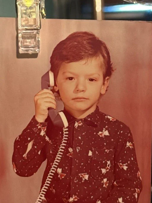 A young boy with brown hair holding a beige telephone receiver to his ear, wearing a dark patterned shirt, standing in front of a wall with electrical outlets.