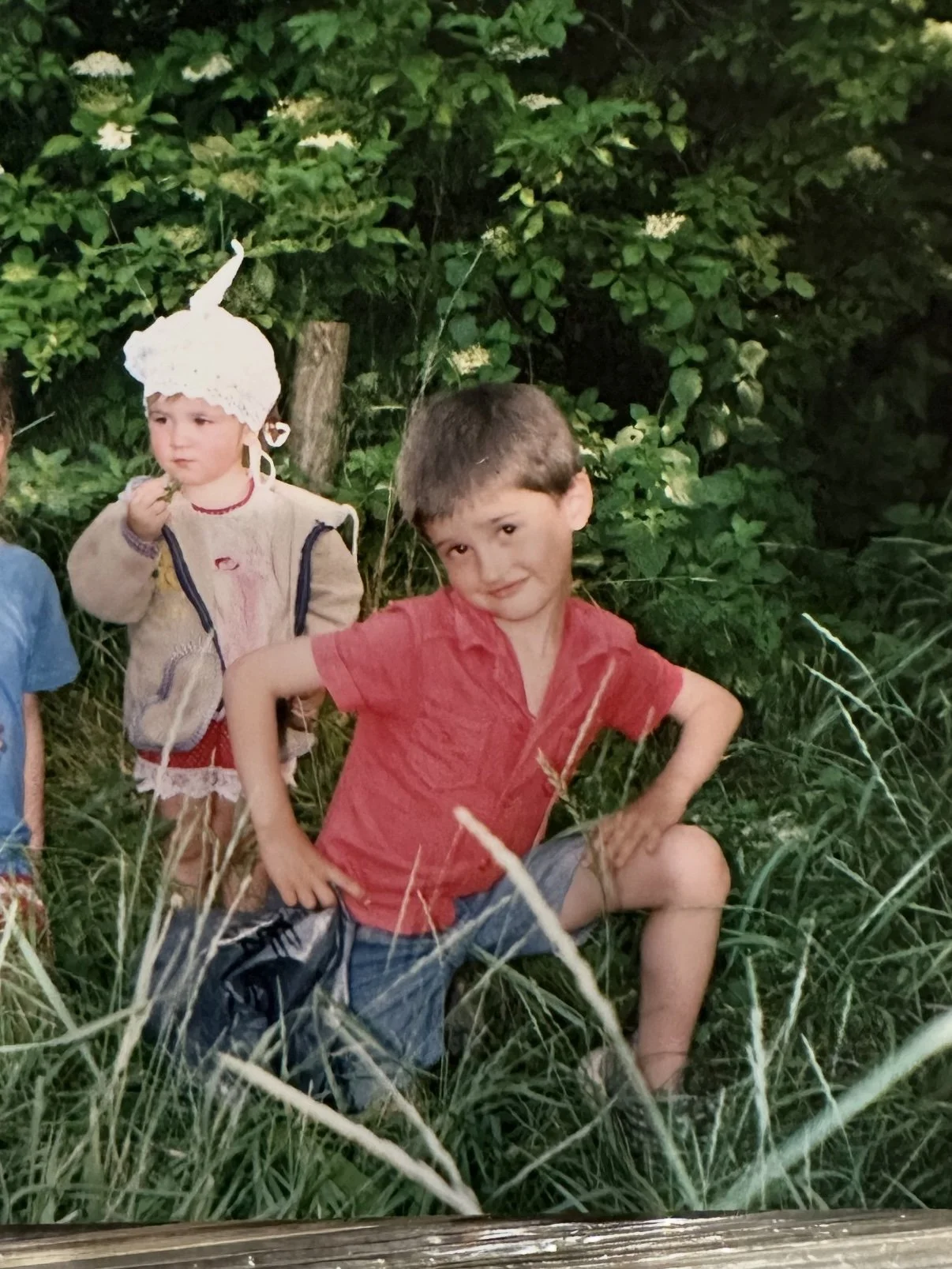Two children outdoors in a grassy area surrounded by green foliage. One boy in a red shirt is sitting on a log with a confident pose, while a girl in a light sweater and a whimsical white hat with a tall, pointy top stands nearby, looking contemplative.