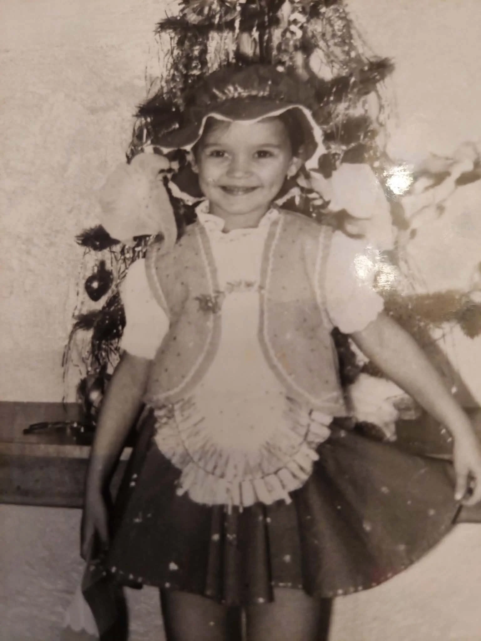 A young girl with a big smile sitting on a chair, wearing a short-sleeved dress with a pleated skirt, and a headband with a decorative pattern, in a room with plain walls.