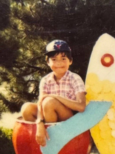 A young boy wearing a baseball cap and a pink plaid shirt, sitting on a colorful, cartoonish airplane-shaped playground structure outdoors with trees in the background.