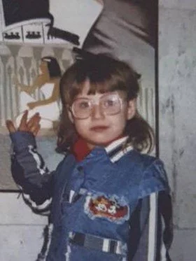 Young girl with glasses wearing a denim jacket with patches, standing indoors near a wall with artwork.