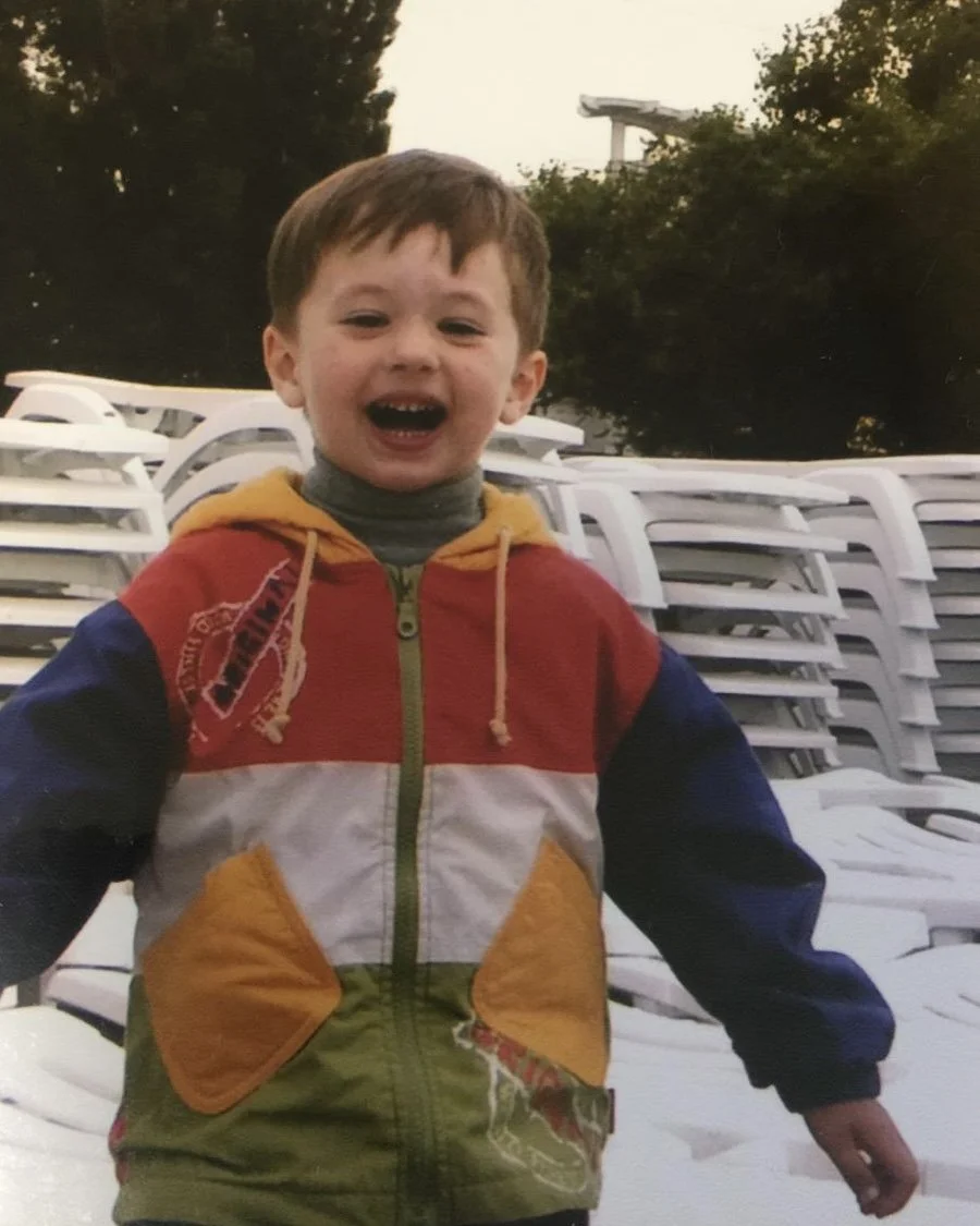 A young boy with brown hair smiling and laughing outdoors, wearing a colorful jacket with yellow, red, blue, and green sections, in front of stack chairs and trees.