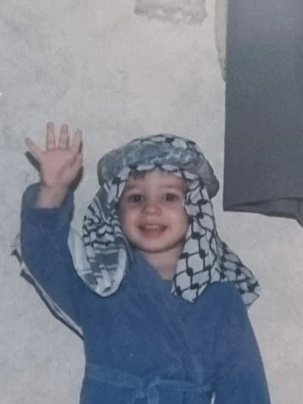 Smiling young child waving, wearing a blue outfit and a patterned headscarf, standing indoors.