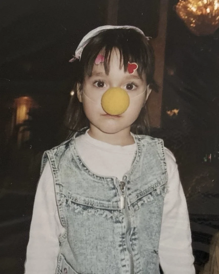 A young girl with brown hair, wearing a denim vest over a white shirt, has a large yellow circular object on her nose and two small red heart-shaped stickers on her forehead.