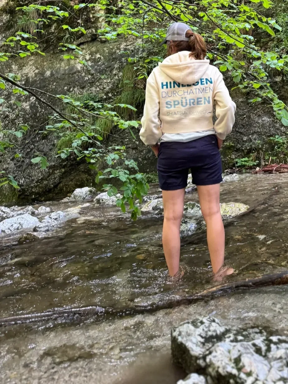 Elisabeth Hollinger – Shiatsu Lilli – steht barfuß im Bach, Hoodie mit Aufschrift Hinlegen Durchatmen Spüren