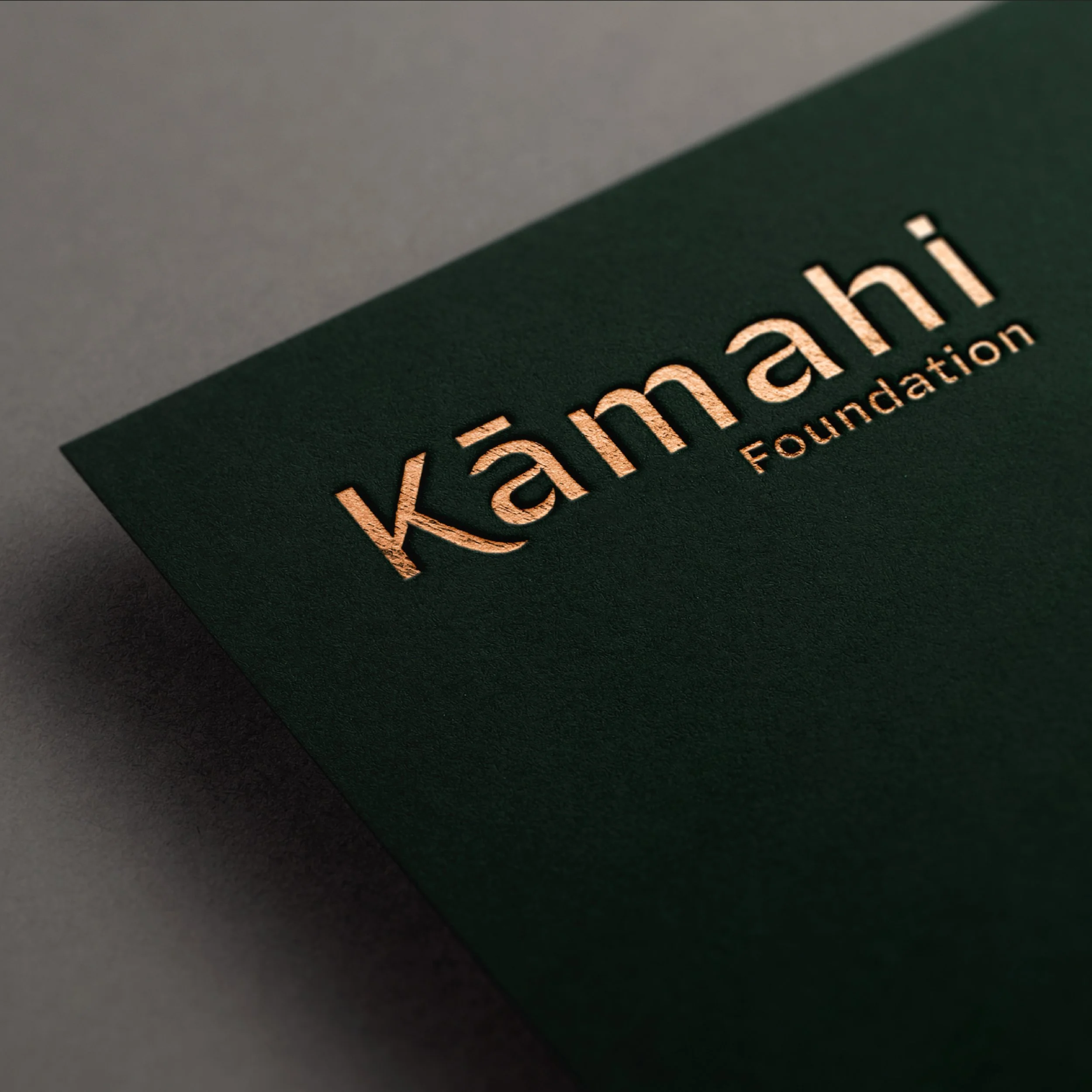 Close-up of a dark green card with the word 'Kāmāhi' and 'Foundation' embossed in copper or bronze in the center.