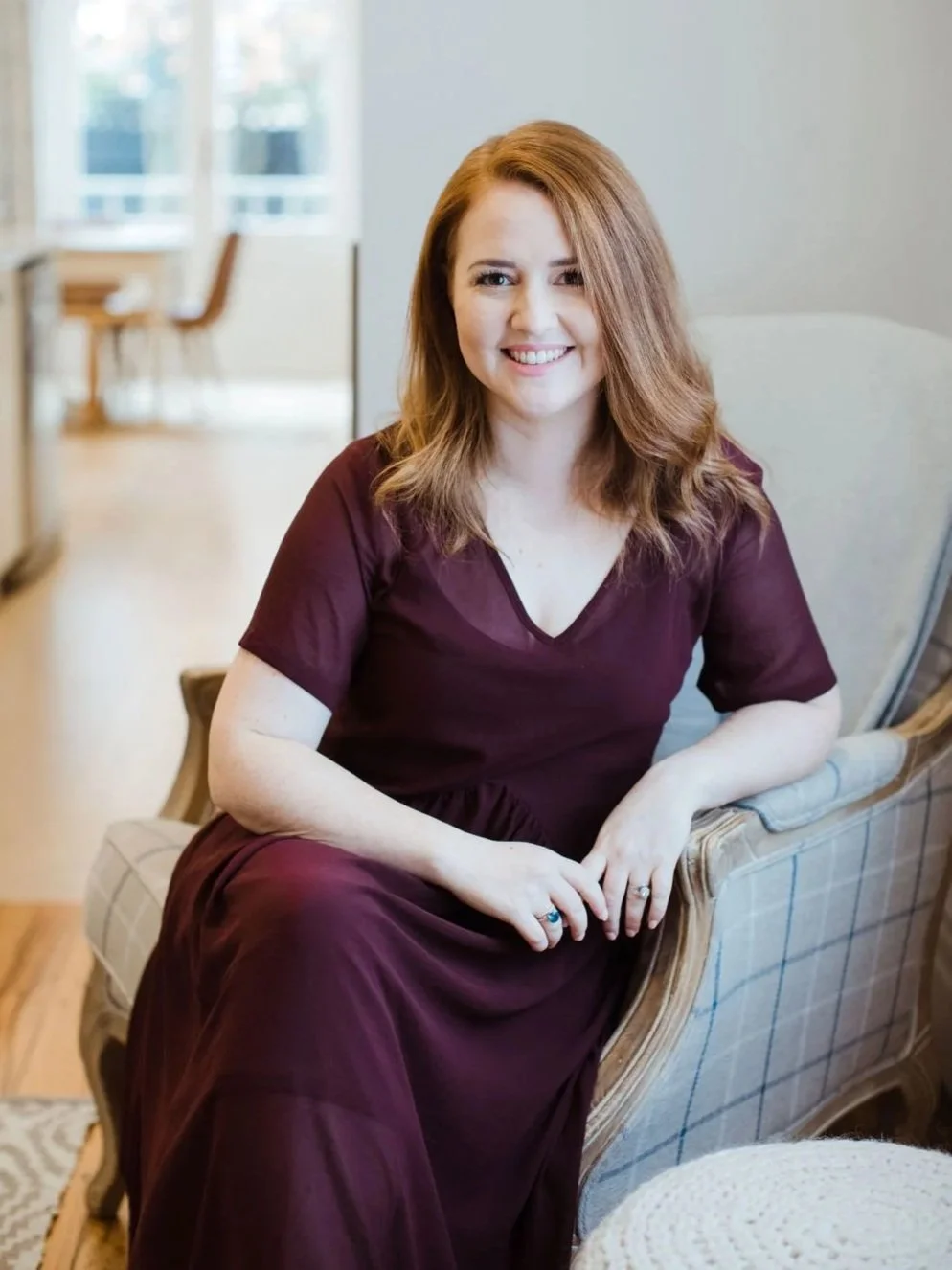 A woman with red hair smiling and sitting on a vintage-style armchair in a cozy, well-lit room.