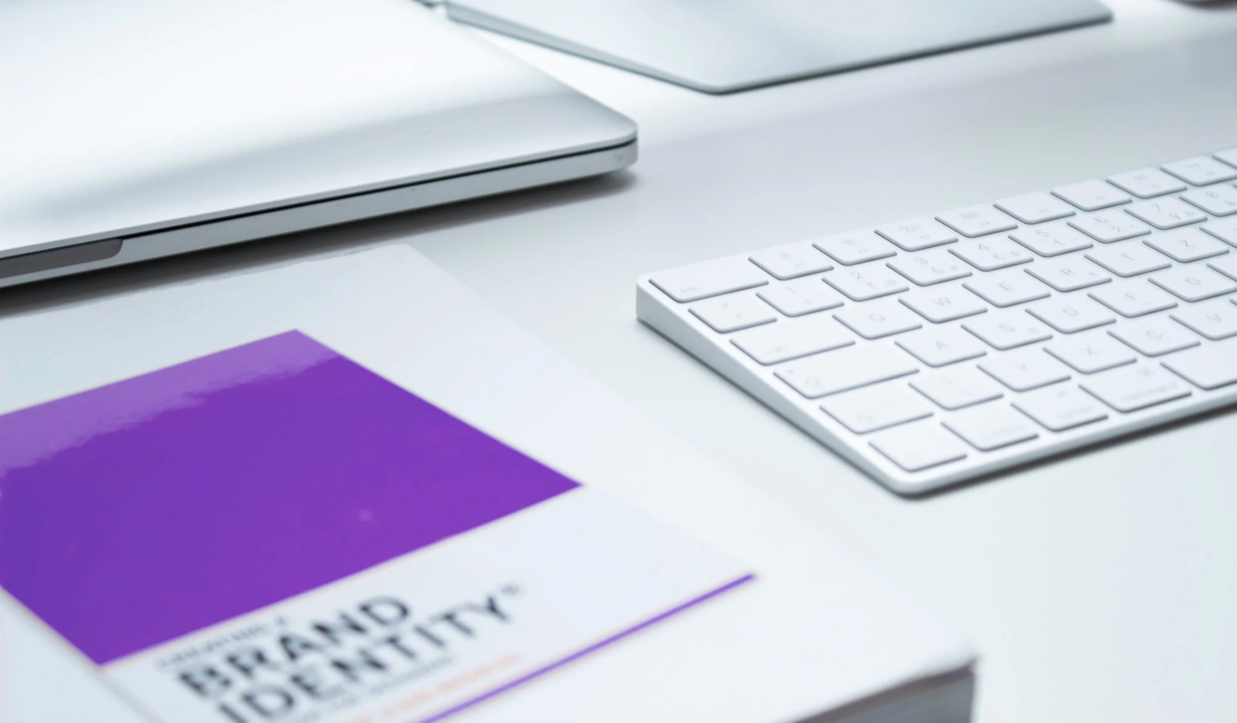 Close-up of a clean workspace with a purple and white book titled 'Brand Identity', a white computer keyboard, and a closed silver laptop on a white desk.