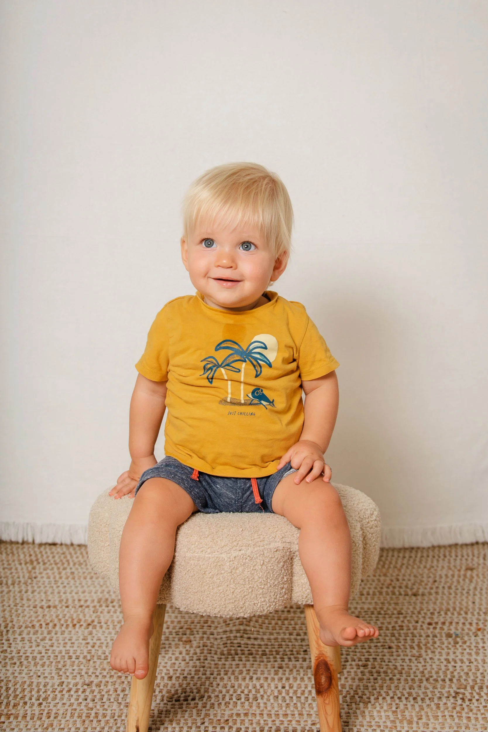 A young blonde-haired boy with blue eyes, wearing a yellow t-shirt with a palm tree design and dark shorts, sitting on a small beige cushioned chair in a room with a neutral white wall and beige carpet.