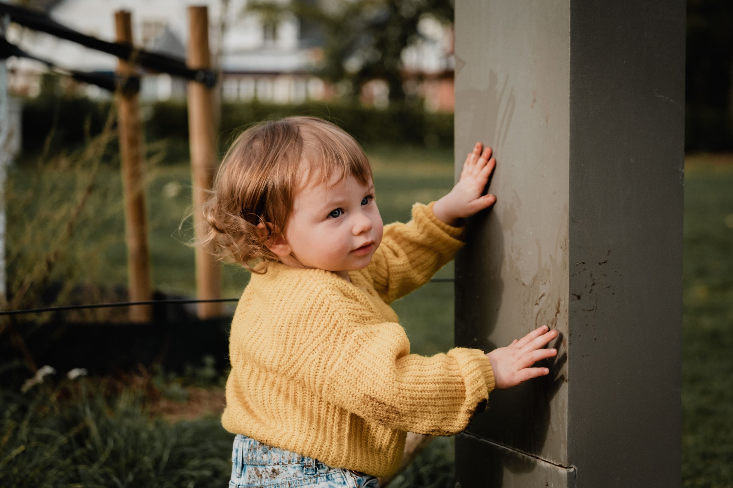 A young girl with curly hair wearing a yellow sweater and jeans, touching a metal sculpture outdoors, with trees and a swing set in the background.