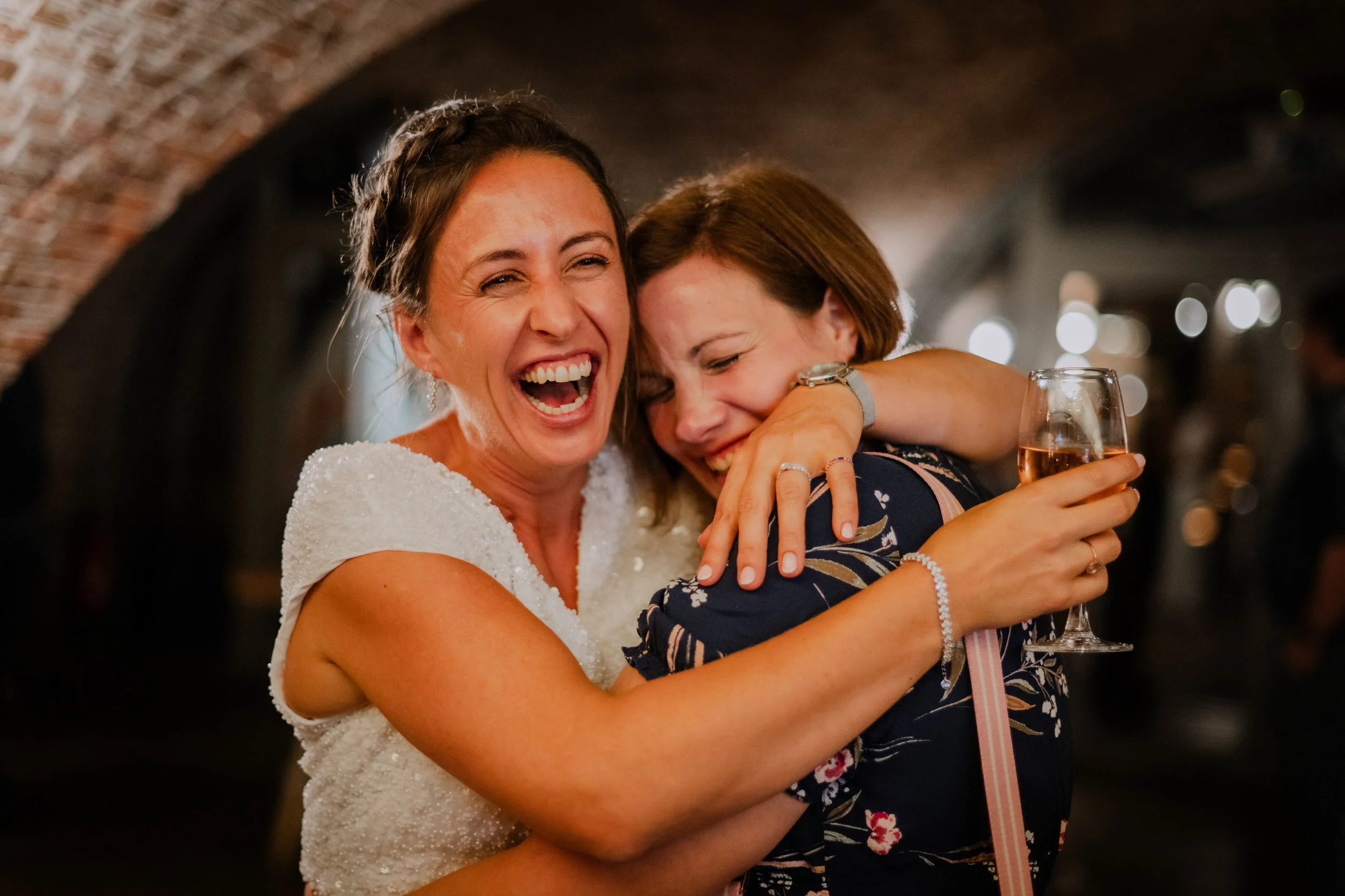 Two women, one in a white dress and the other in a dark floral dress, hugging and laughing at a celebration or party, with a wine glass in hand, in a warmly lit brick-ceiling venue.