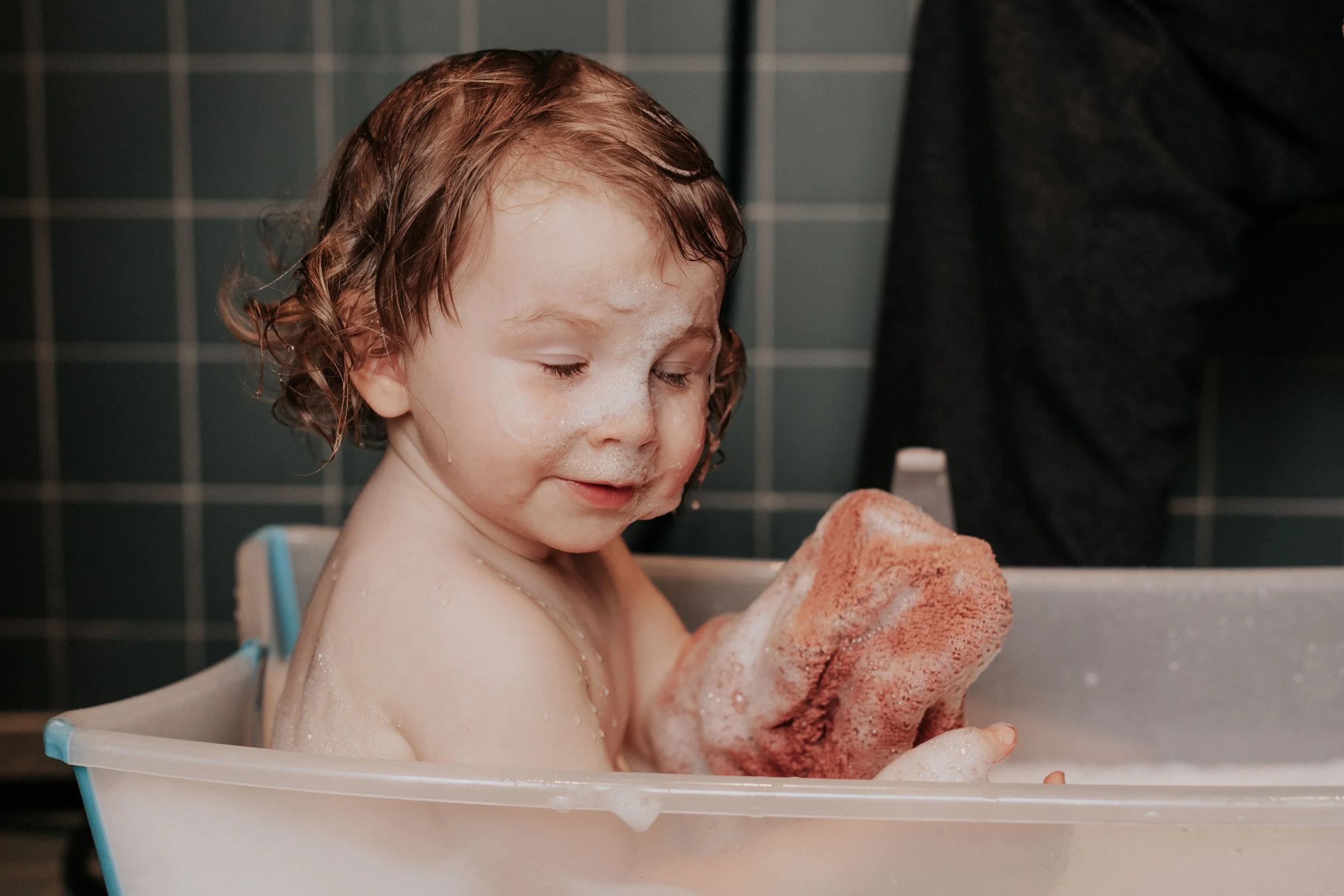 A young child with wet hair and soap suds on their face, sitting in a small tub, holding a sponge, during bath time.