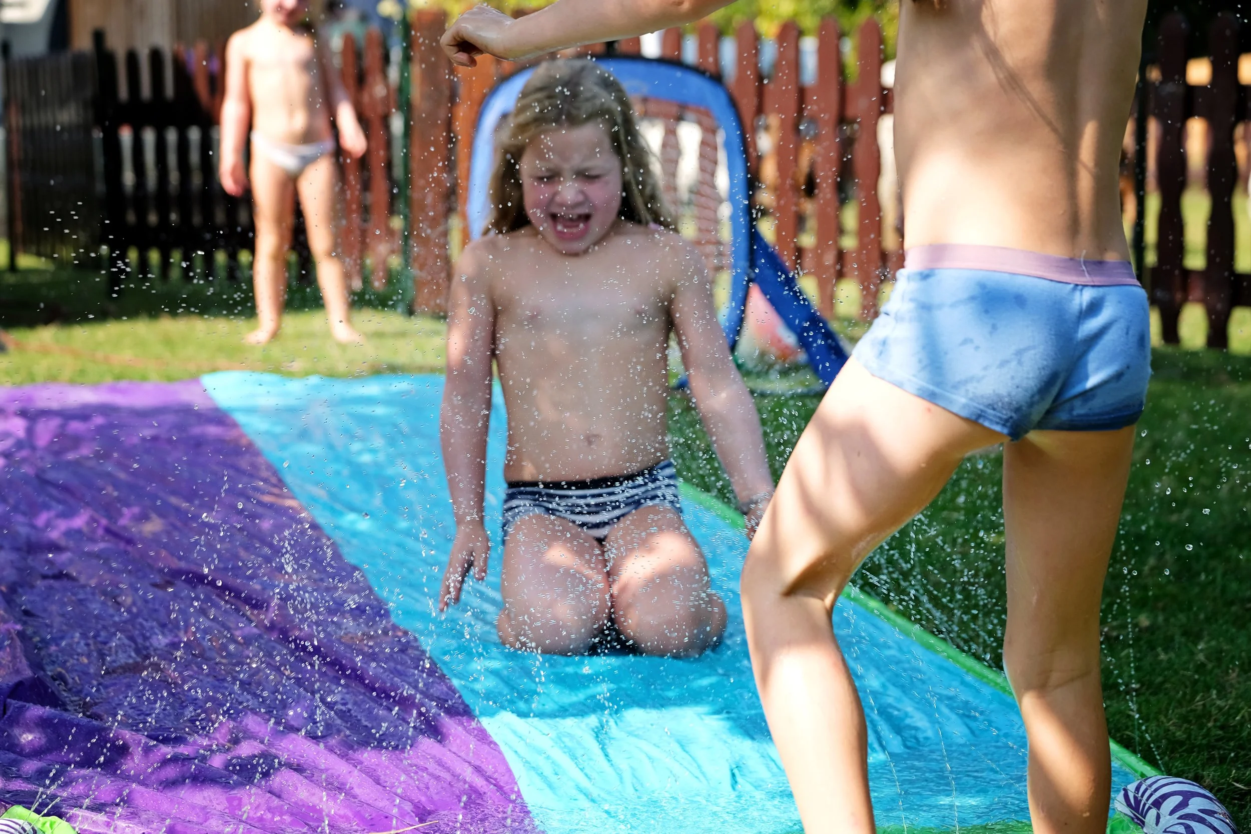 Children playing on a slip 'n slide in a backyard, with one child mid-slide and others in the background.