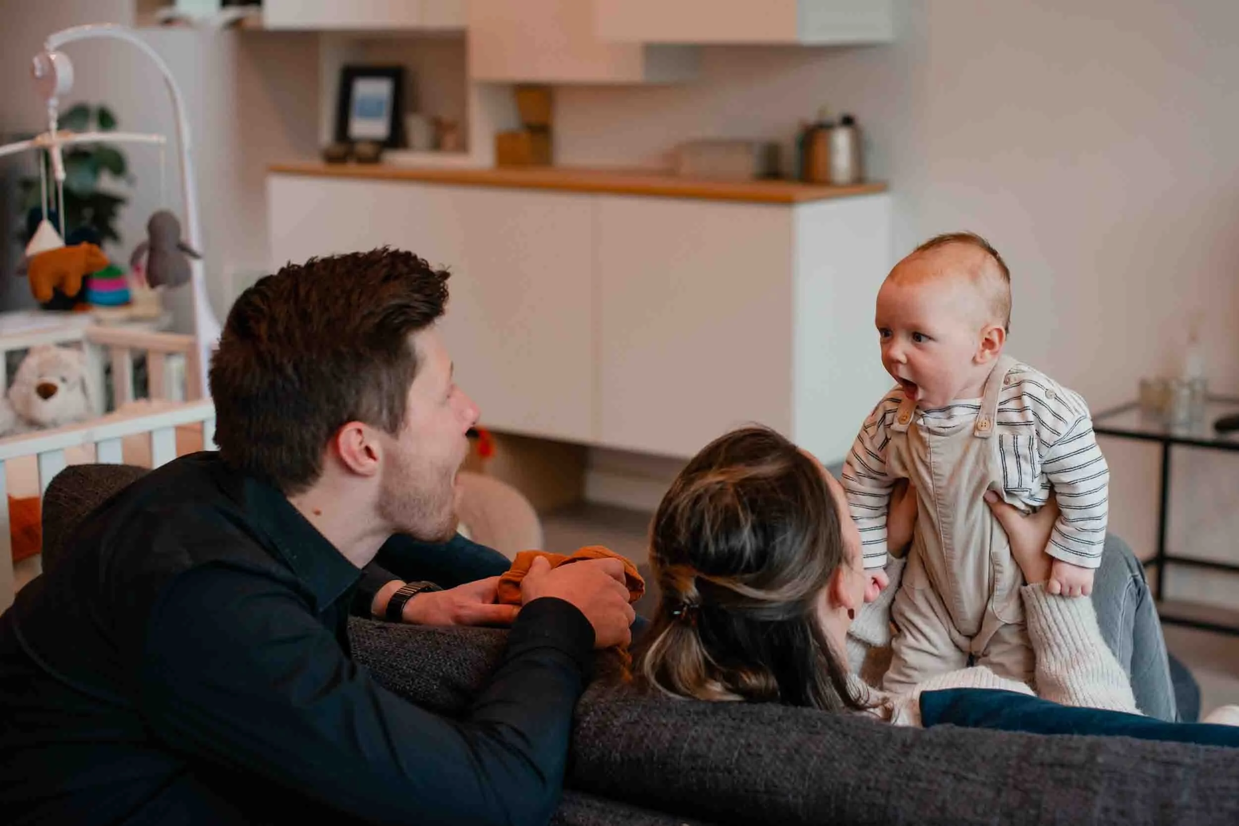 A group of adults and children enjoying a playful moment together in a living room. A baby with a striped shirt is being lifted up by an adult, and other people are gathered around, smiling and interacting.