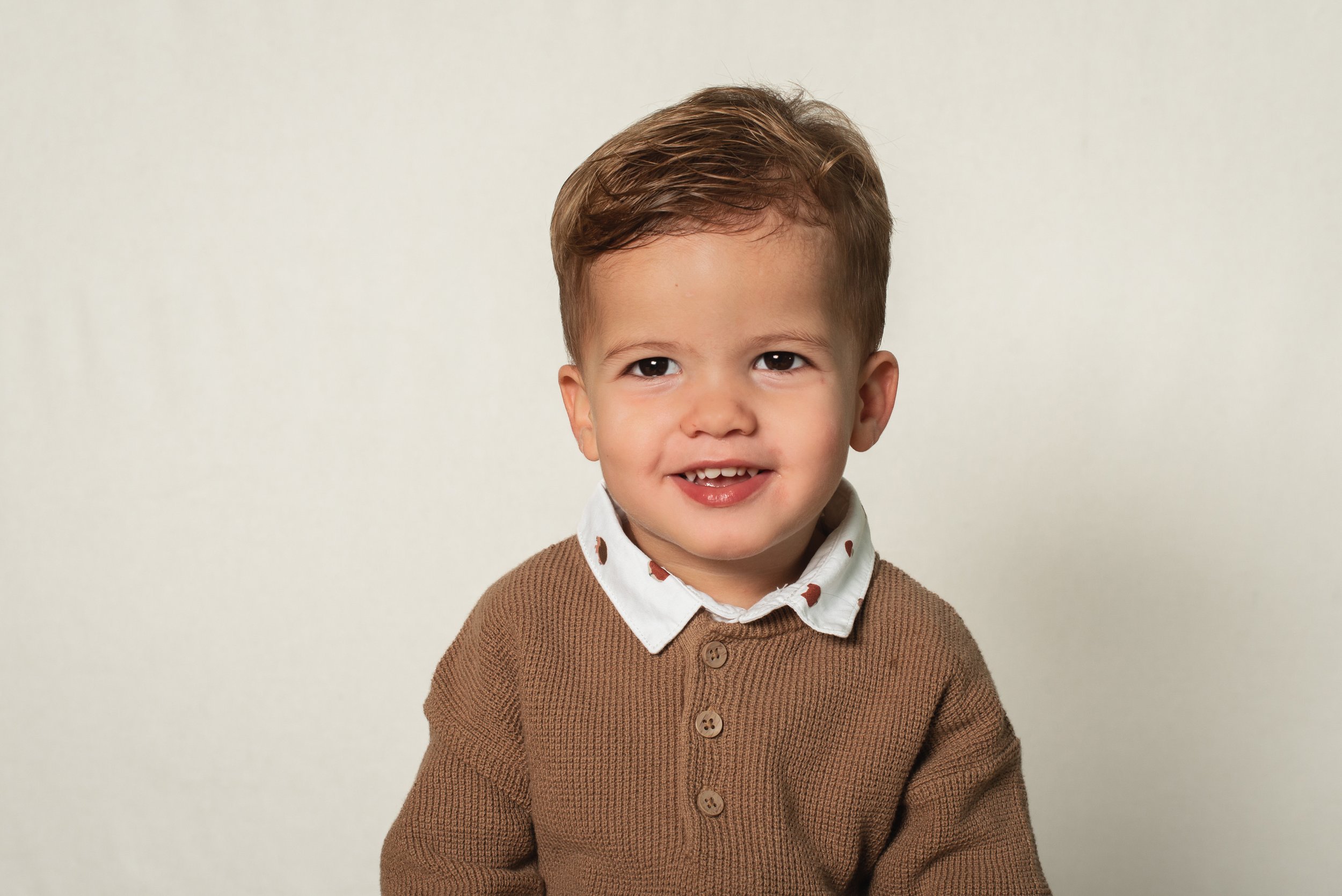 A young boy with brown hair and brown eyes smiling, wearing a brown sweater over a collared shirt with polka dots, standing against a plain light background.