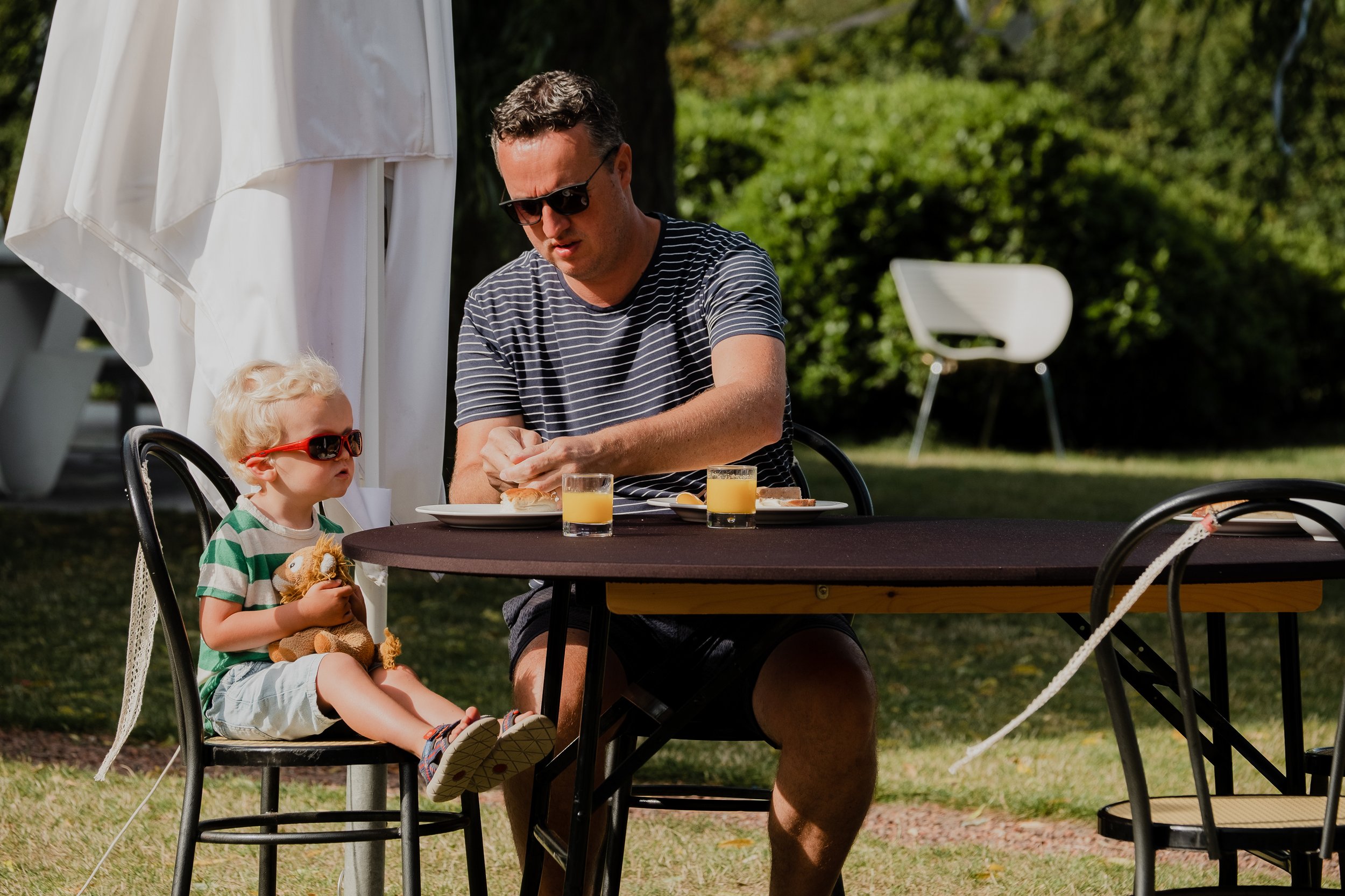 A man and a young child sitting at an outdoor table, having breakfast. The man is wearing sunglasses and a striped shirt, the child has blonde hair, red sunglasses, and is holding a teddy bear. There is a white flag or curtain in the background, and 