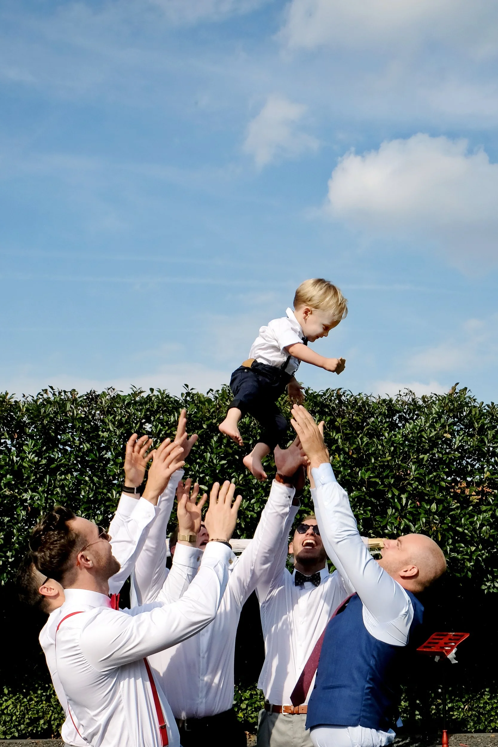 A group of men in formal attire, some with sunglasses, are lifting a young boy in a white shirt and black pants into the air outdoors. The boy is smiling, and the men are cheering with raised hands. There are green bushes and a blue sky with clouds i