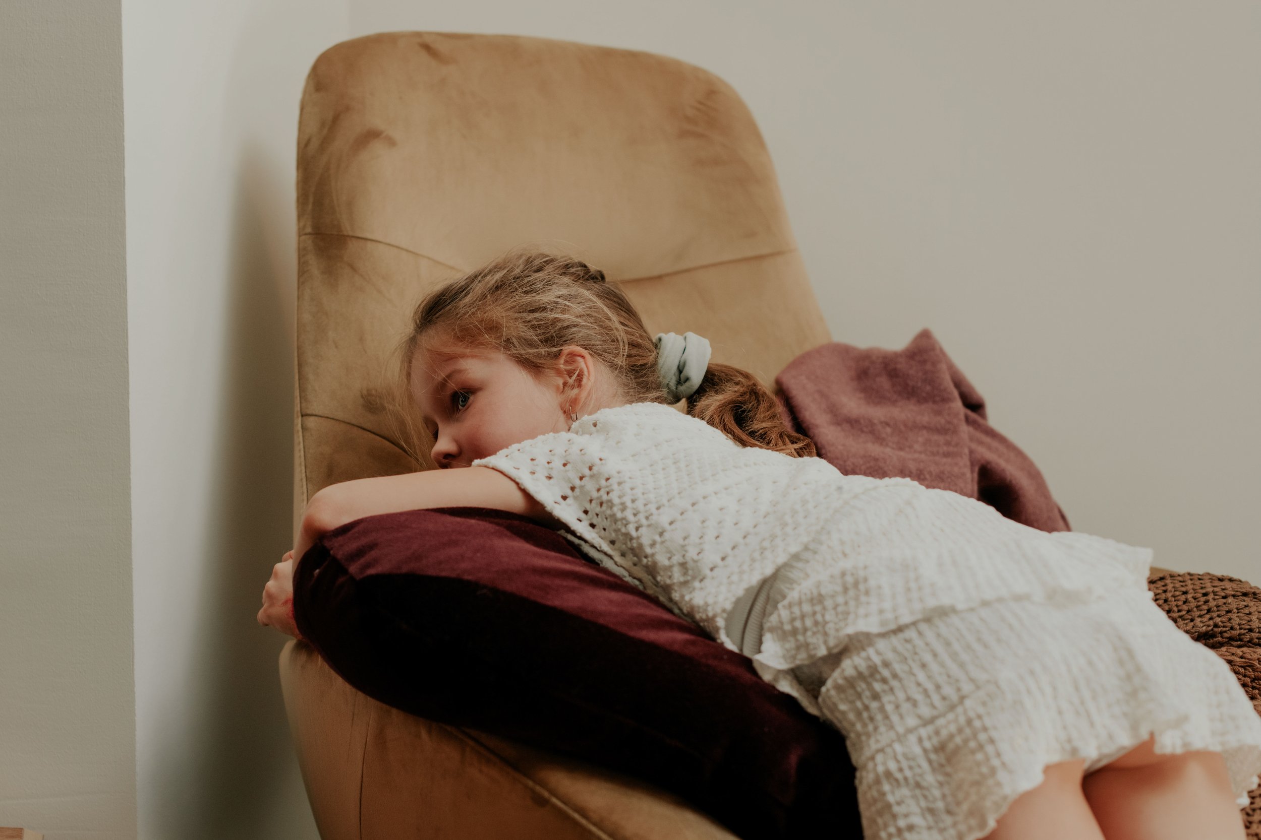 A young girl with light brown hair lying on her stomach on a bed, resting her head on her arms, with a beige headband and a white textured top. She is next to a brown blanket and a wicker basket, with a plain wall and headboard behind her.