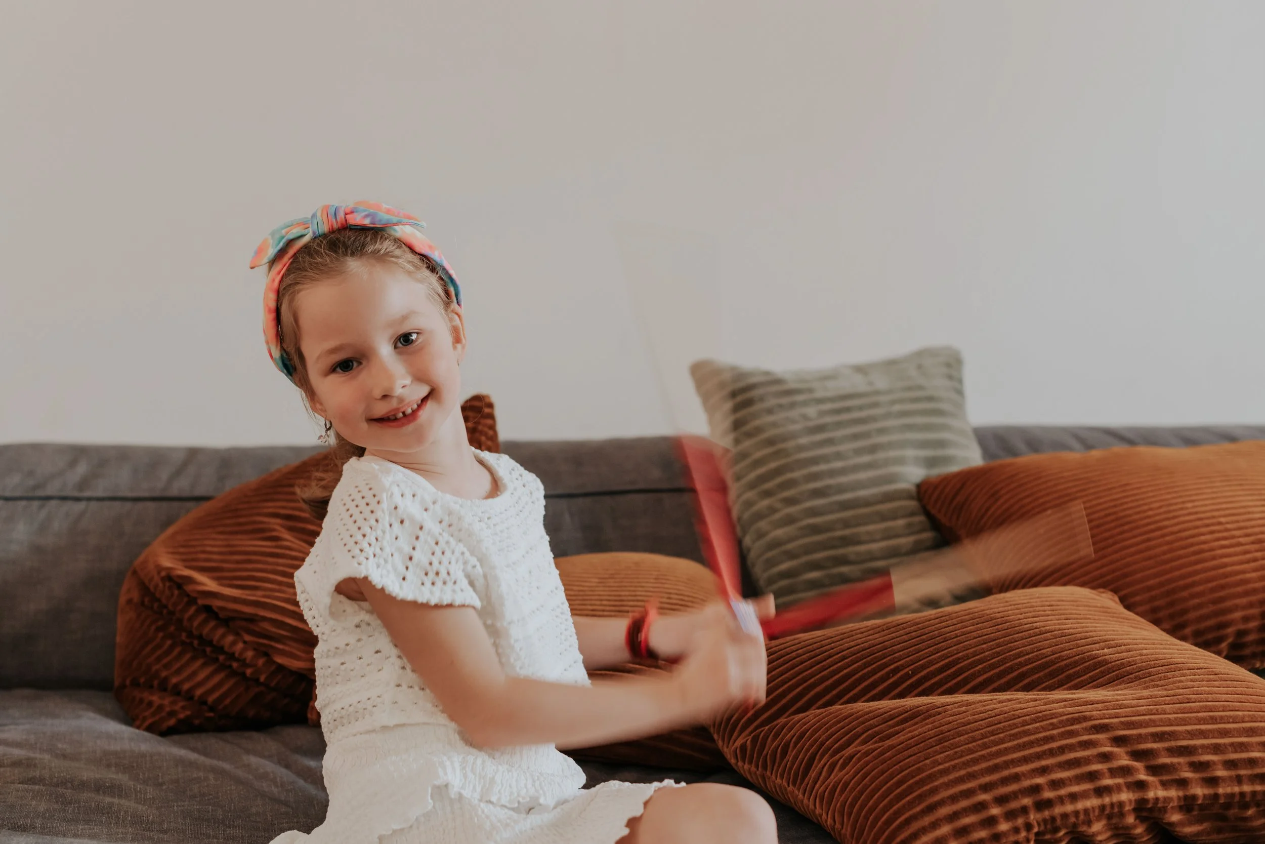 A young girl with a colorful headscarf smiling at the camera, sitting on a sofa with brown and striped pillows against a plain white wall.