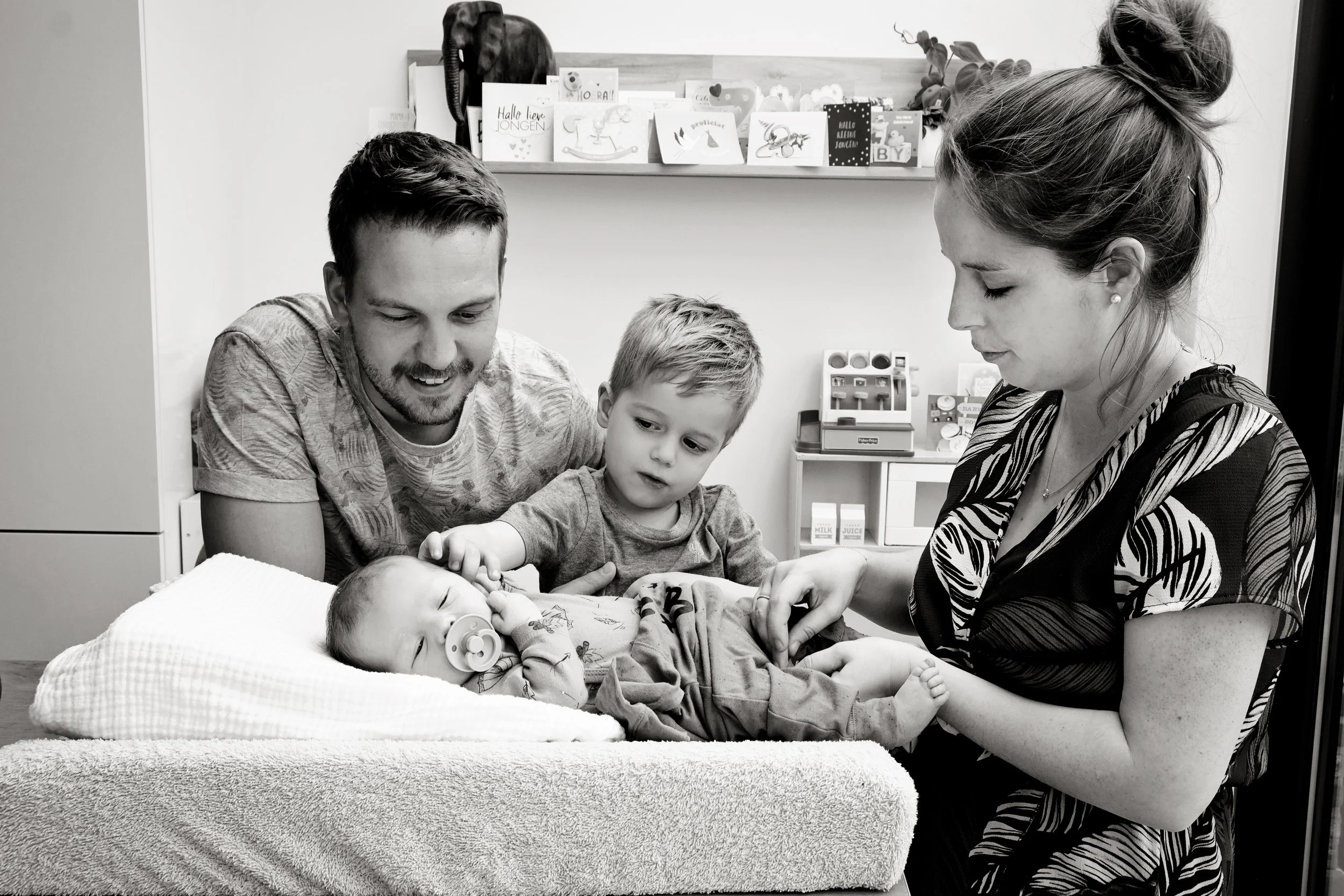 A family including a man, a woman, and two young children are gathered around a hospital bed with a baby, in a medical or hospital room decorated with greeting cards and toys. The woman appears to be Mother’s Day or birthday celebrating.