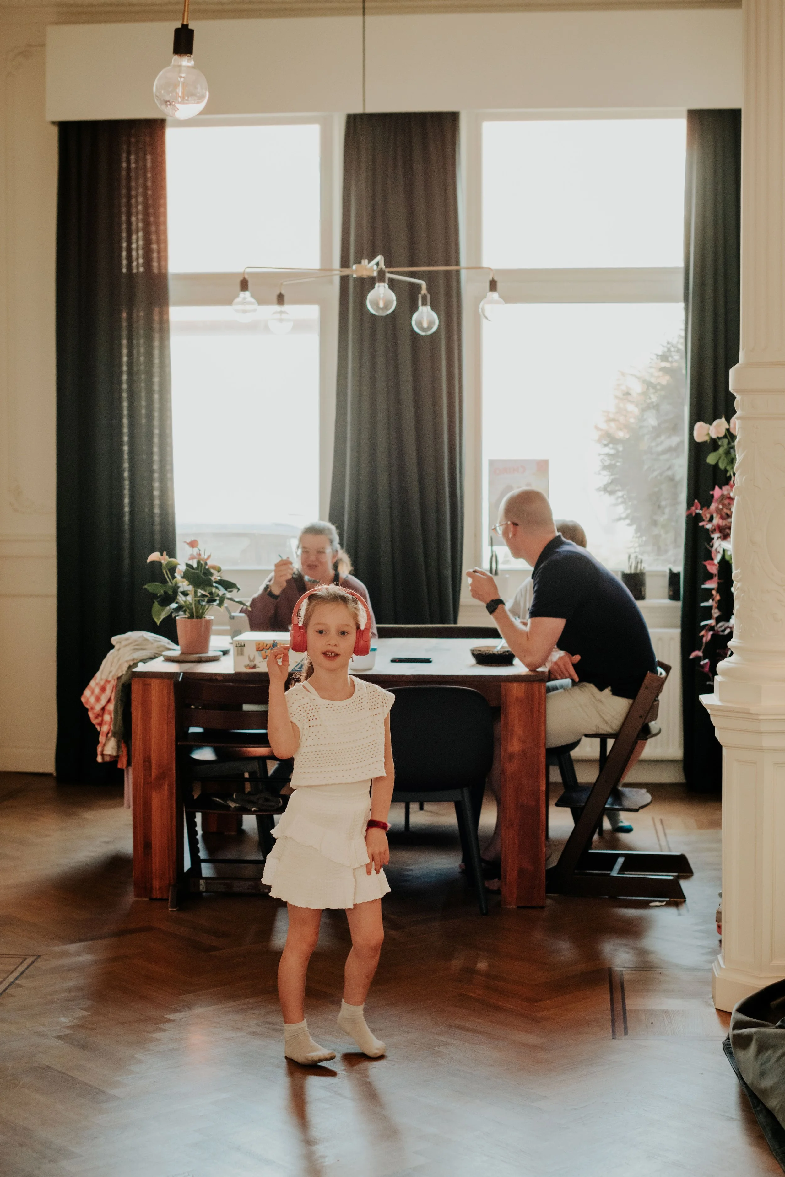 A young girl with red headphones, wearing a white outfit, standing in a room with wooden floors, with people dining in the background near large windows with curtains.