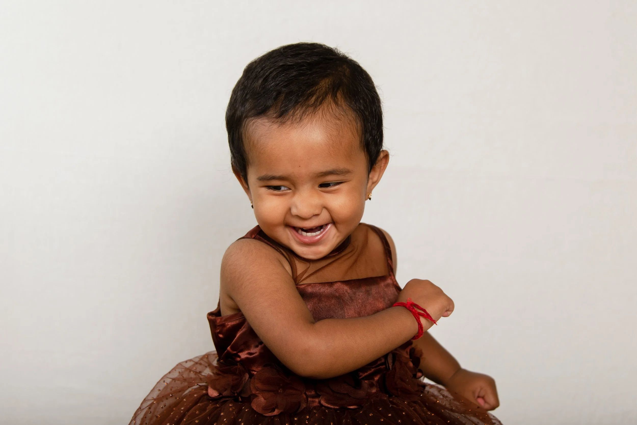 Young girl with short dark hair, wearing a brown dress with floral patterns and a red bracelet, smiling and looking to her right against a plain white background.