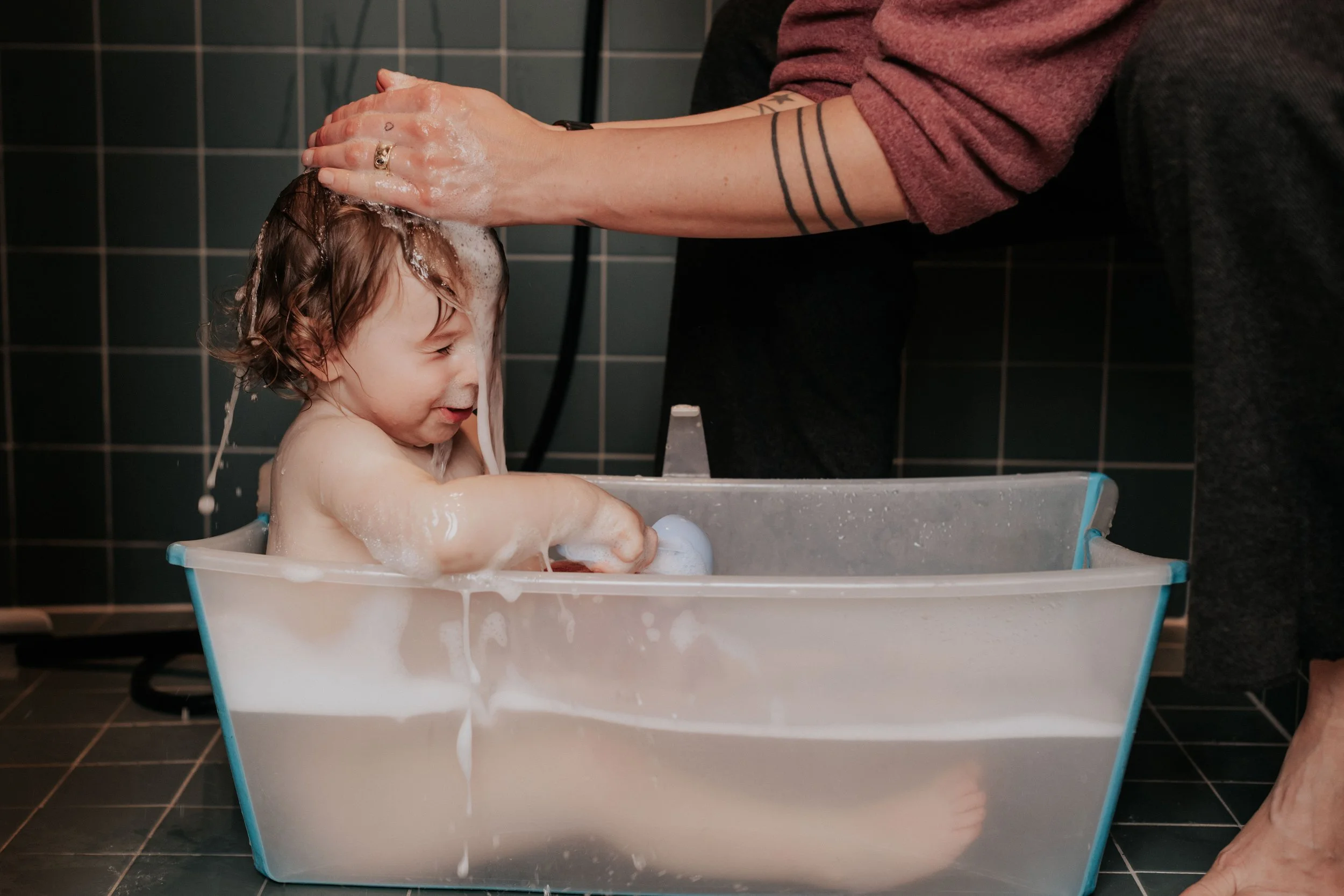A young child taking a bath in a plastic tub while an adult nearby washes their hair with shampoo and water in a tiled bathroom.