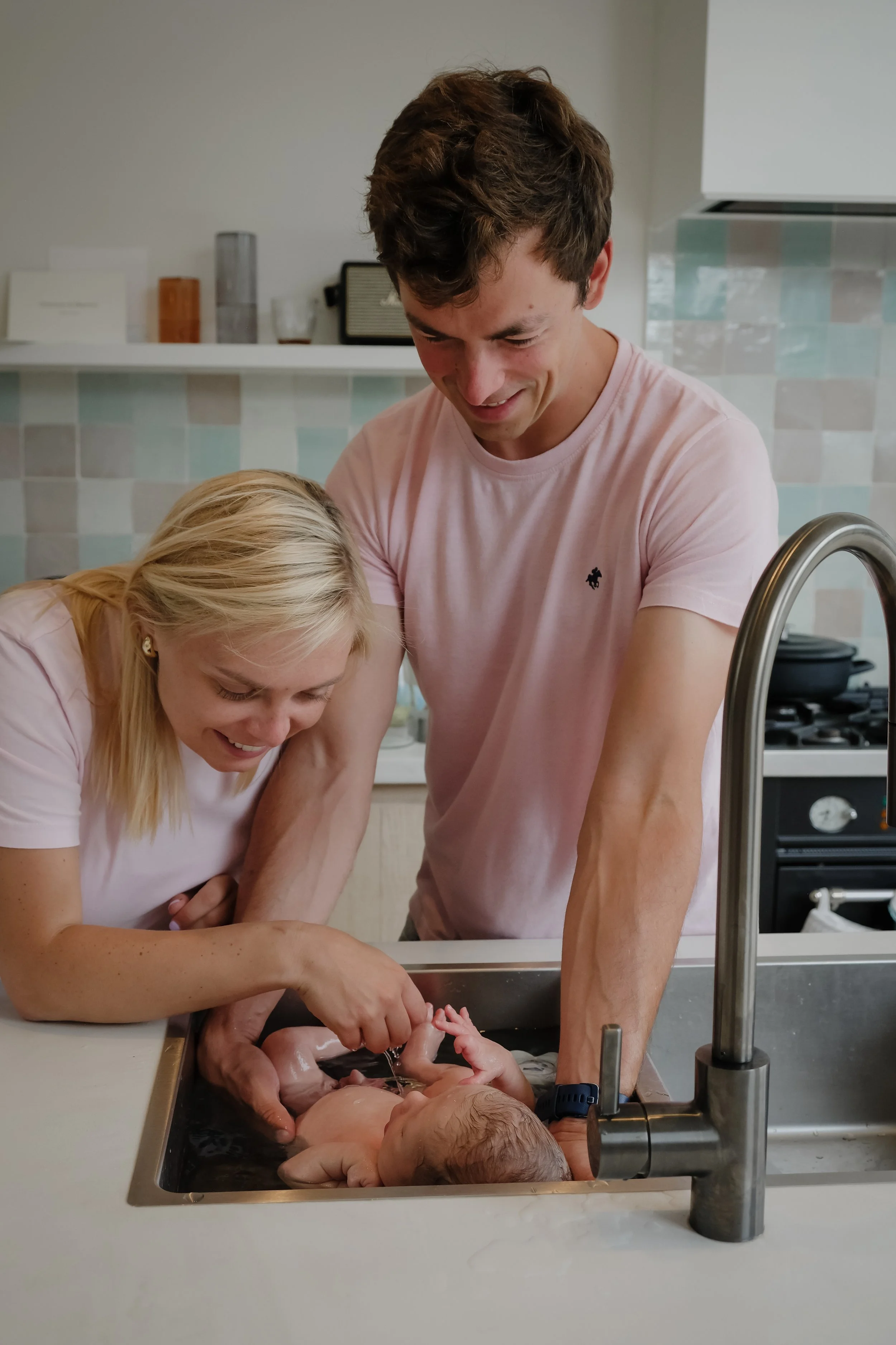 A man and woman in a kitchen giving a newborn baby a bath in the sink.