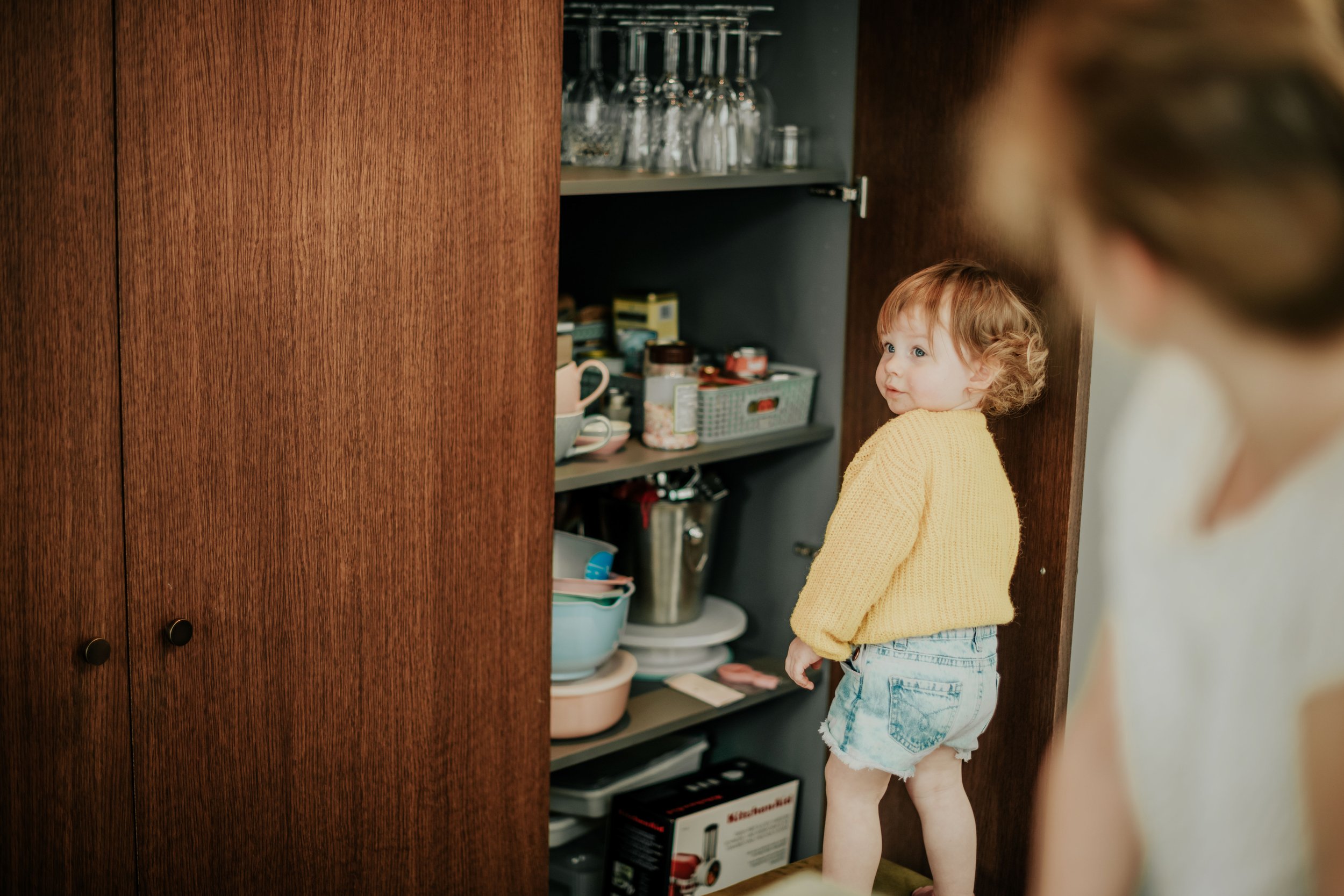 Young girl with curly red hair wearing a yellow sweater and denim shorts standing in a kitchen near a wooden cabinet, looking inside a shelf with glasses and kitchen supplies.