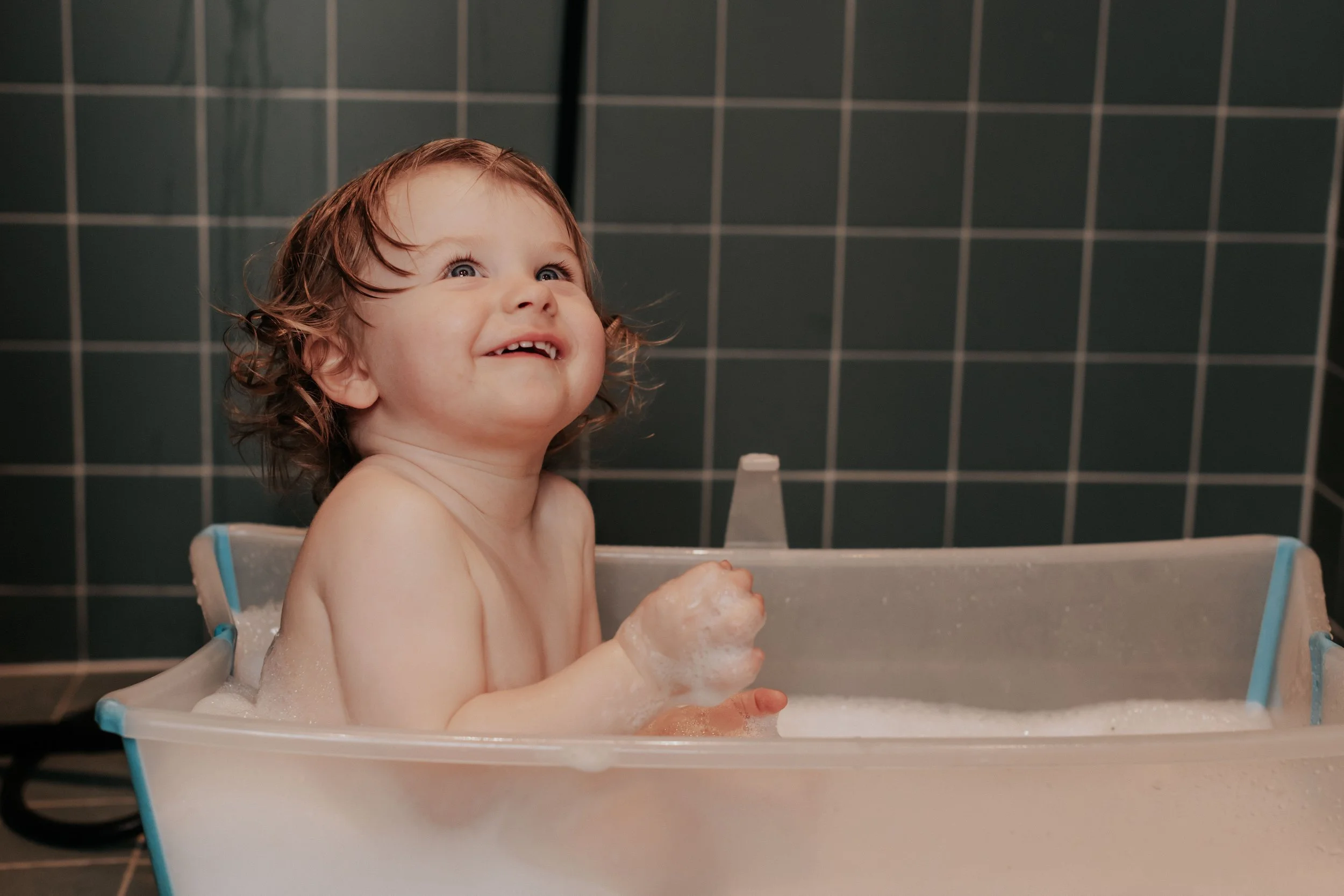 A young child with wet, curly hair smiling while bathing in a plastic tub, surrounded by dark tiled walls.