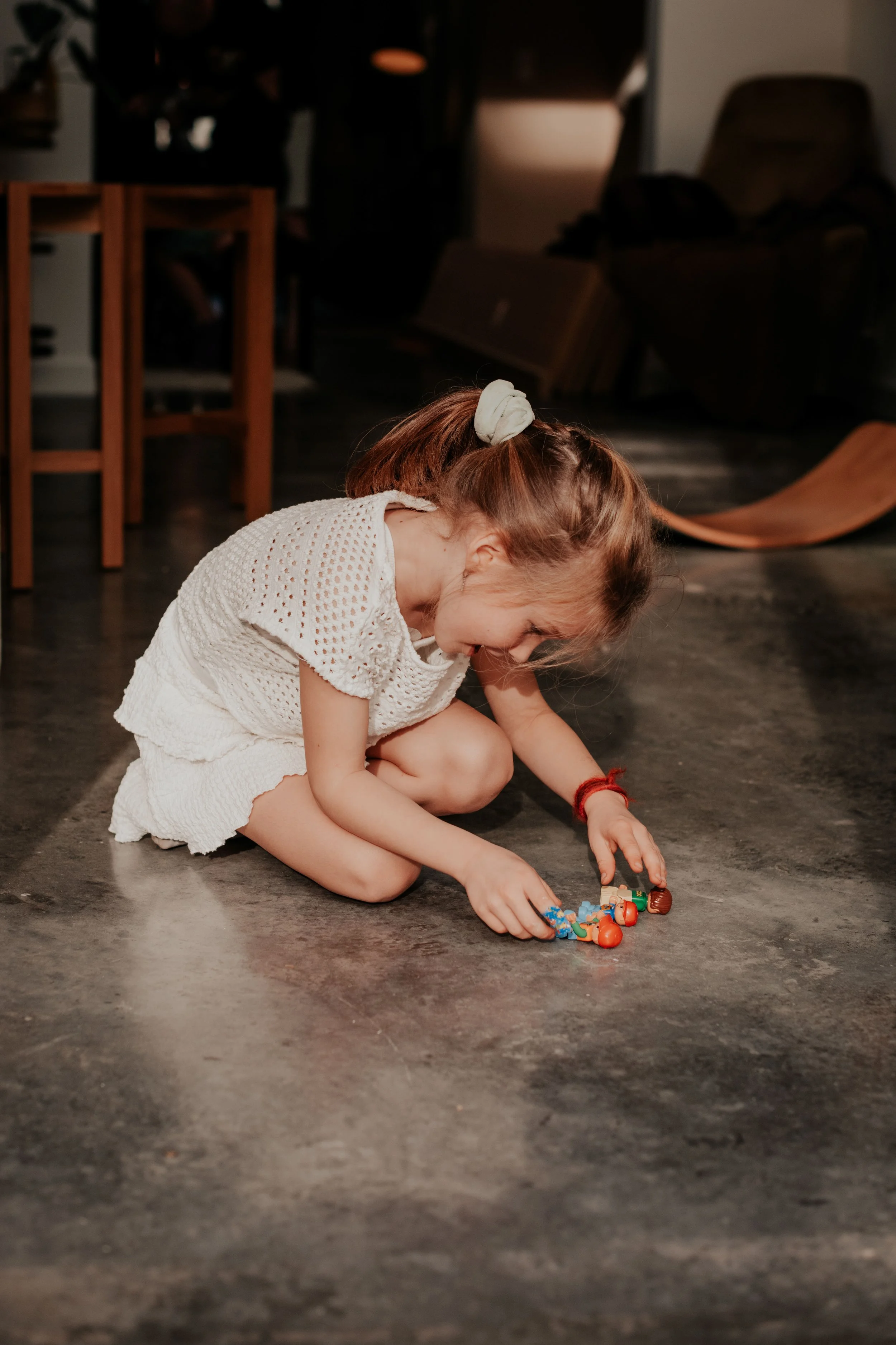 A girl with light brown hair in a ponytail, wearing a white dress, is sitting on the floor and playing with toy cars. The background features wooden chairs and a bench, with a dark, indoor setting.