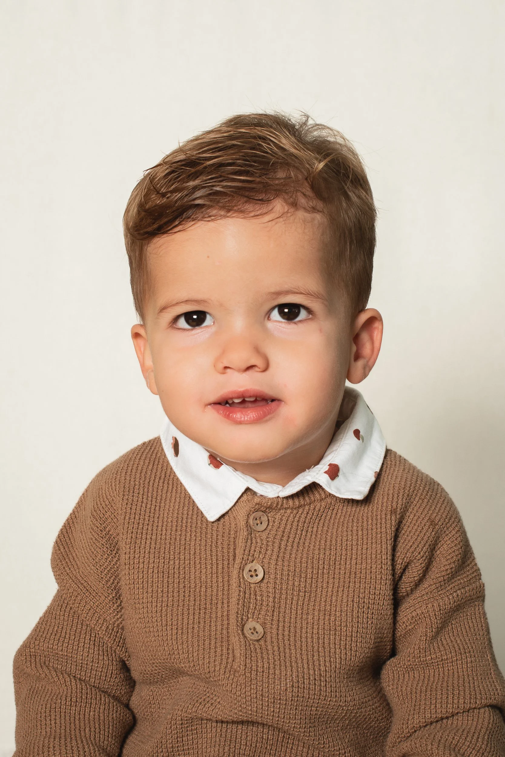 Close-up of a young boy with light brown hair, dark eyes, and fair skin, wearing a white collared shirt with brown spots, layered with a brown knitted cardigan, against a plain white background.