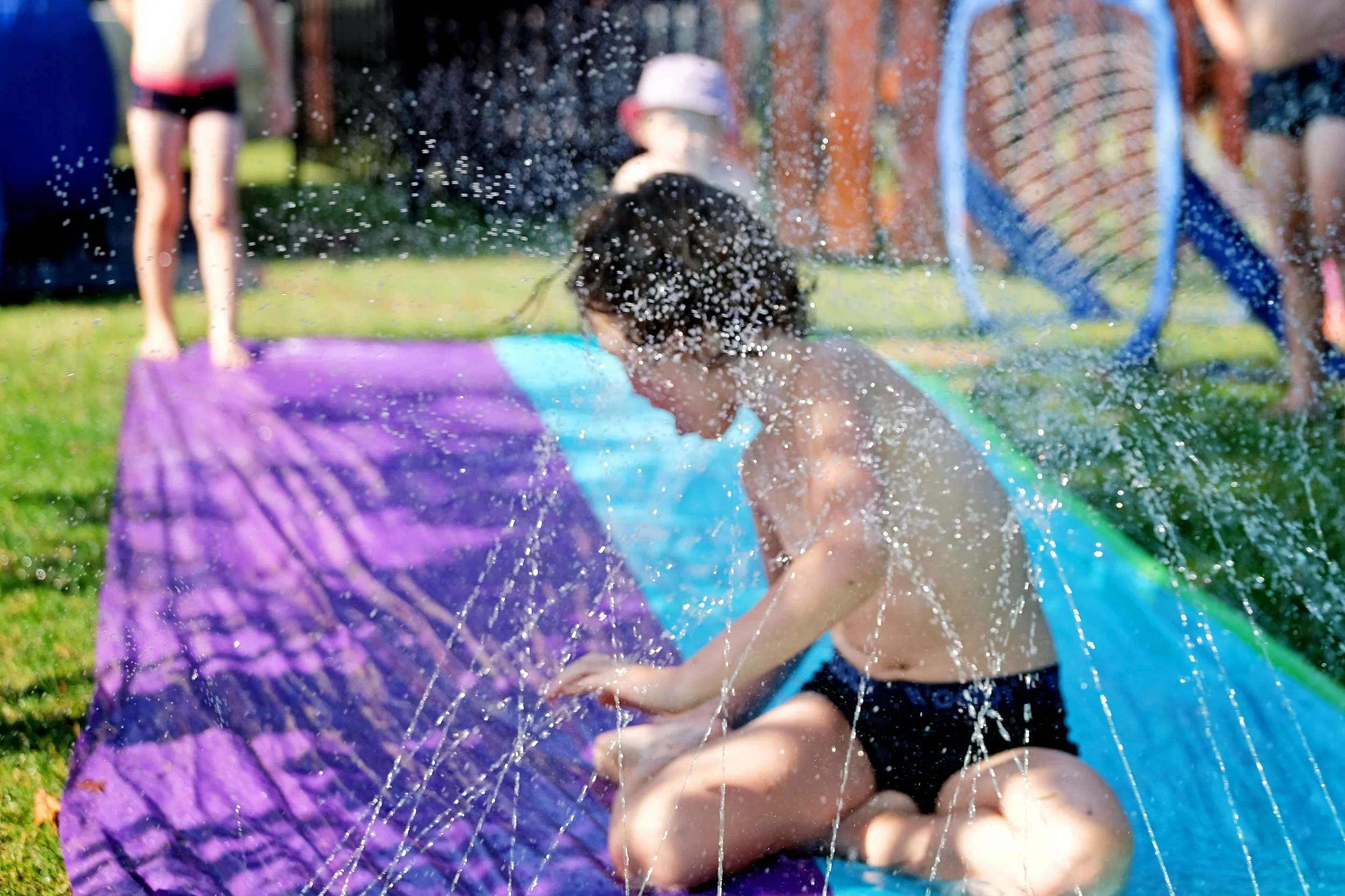 A child playing on a water slide outdoors, with other children in the background, water splashing around, on a sunny day.